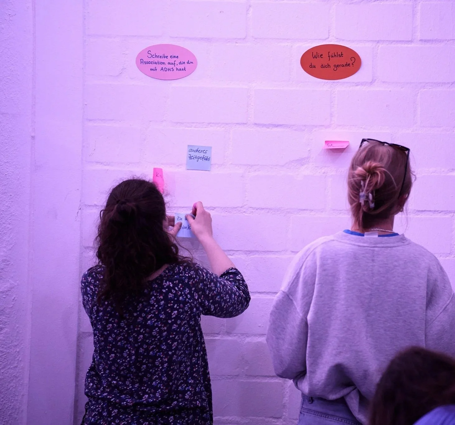 Zwei Frauen schreiben auf Post-it-Zettel, die sie an eine weiße Ziegelwand mit Fragen kleben, in einem Raum mit pinker Beleuchtung.