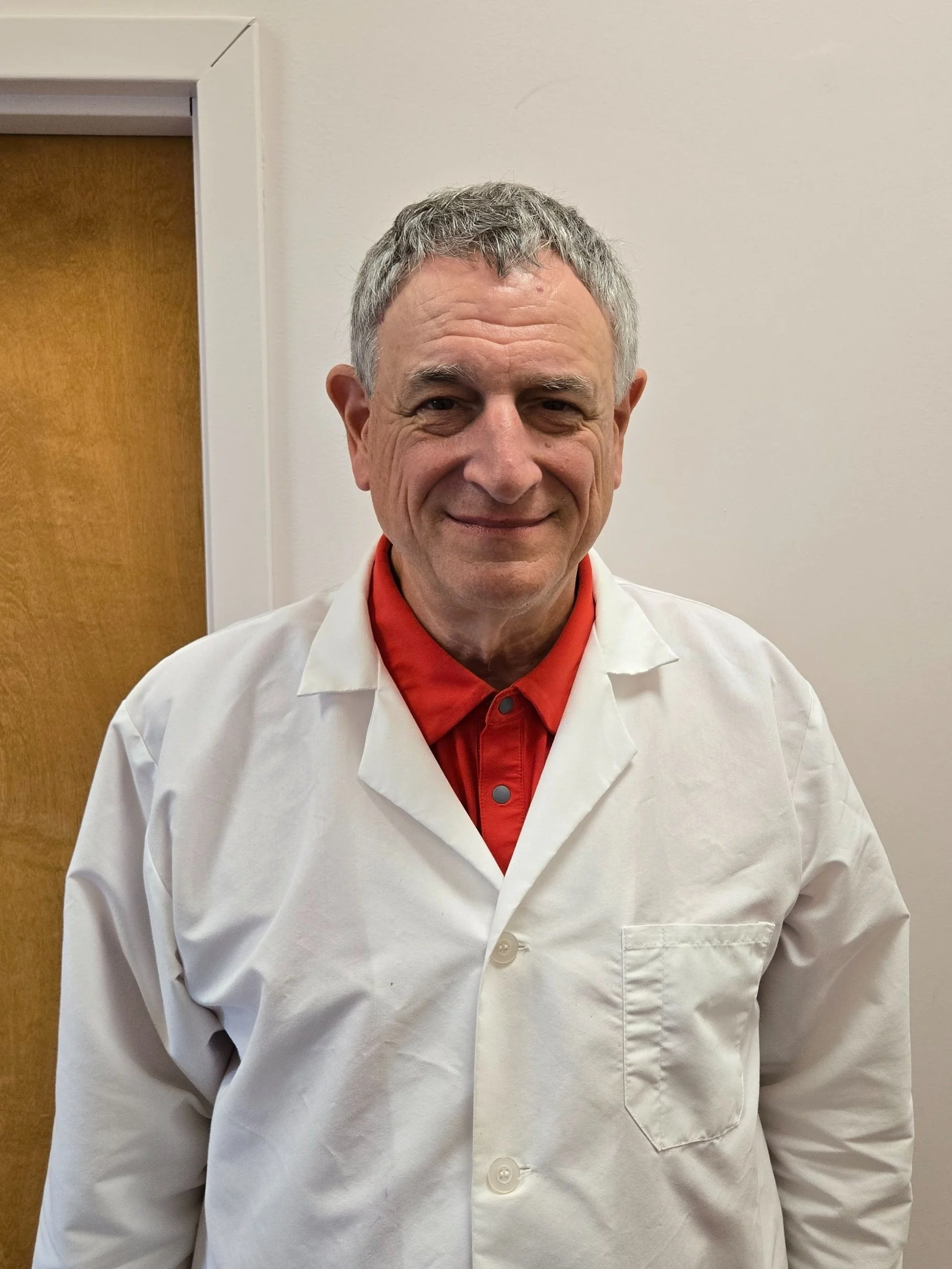 A middle-aged man with short gray hair, smiling, wearing a white lab coat over a red shirt, standing indoors against a plain wall with a wooden door partially visible in the background.