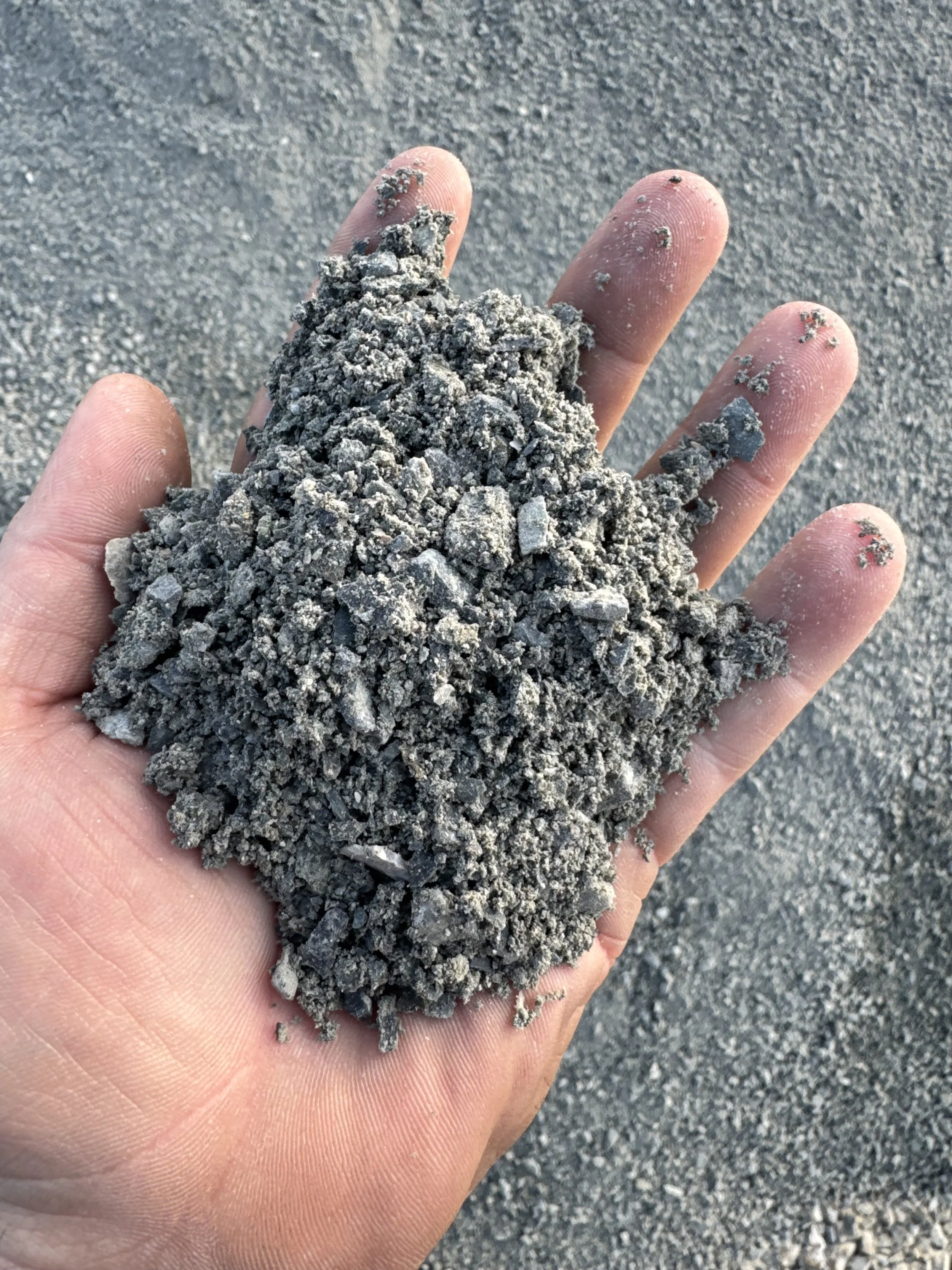 A person's hand holding a pile of gray sand with a rough texture, with a sandy background.