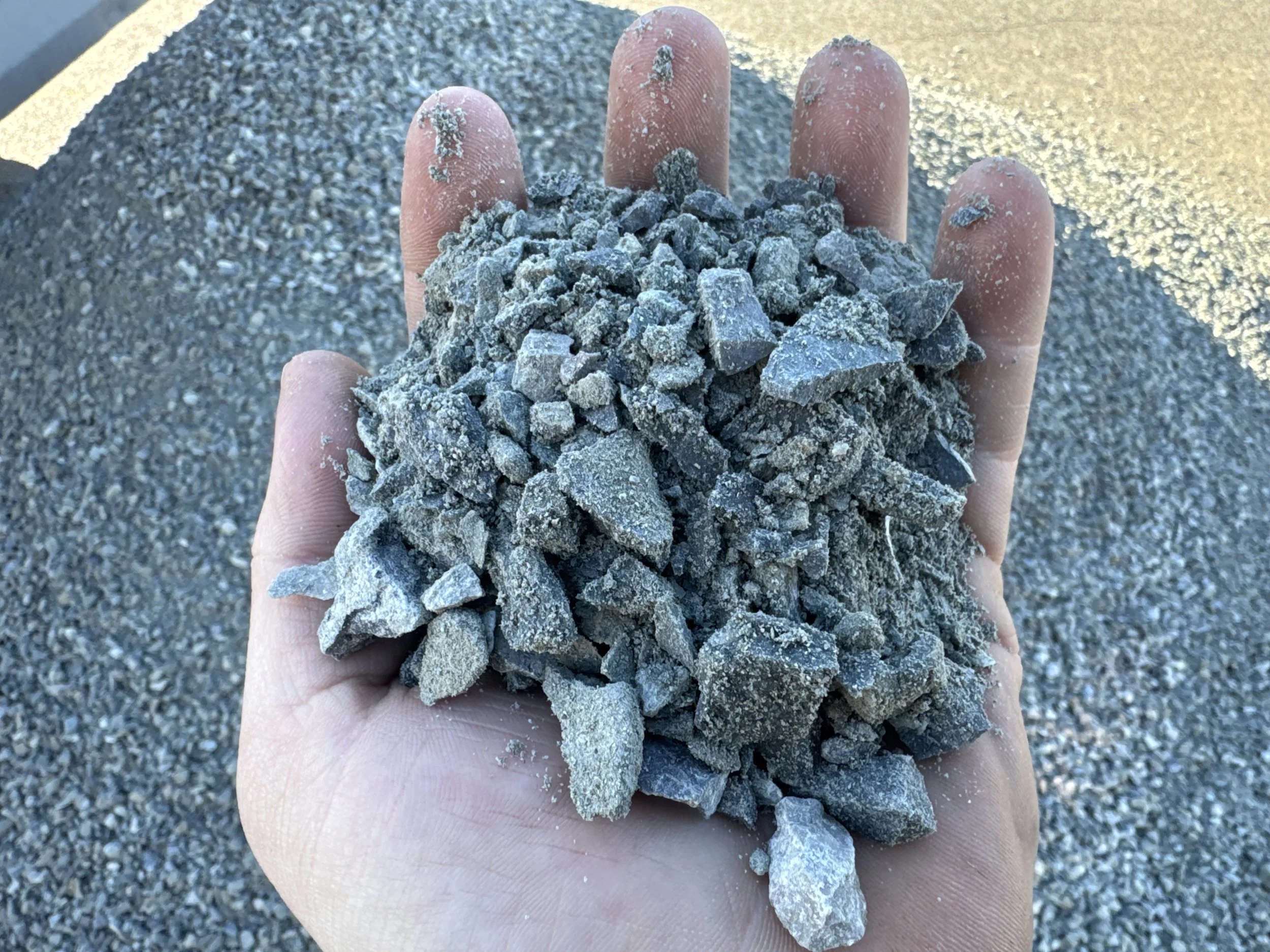 A person's hand holding a pile of small, irregular gray gravel stones outdoors with a large pile of gravel in the background.