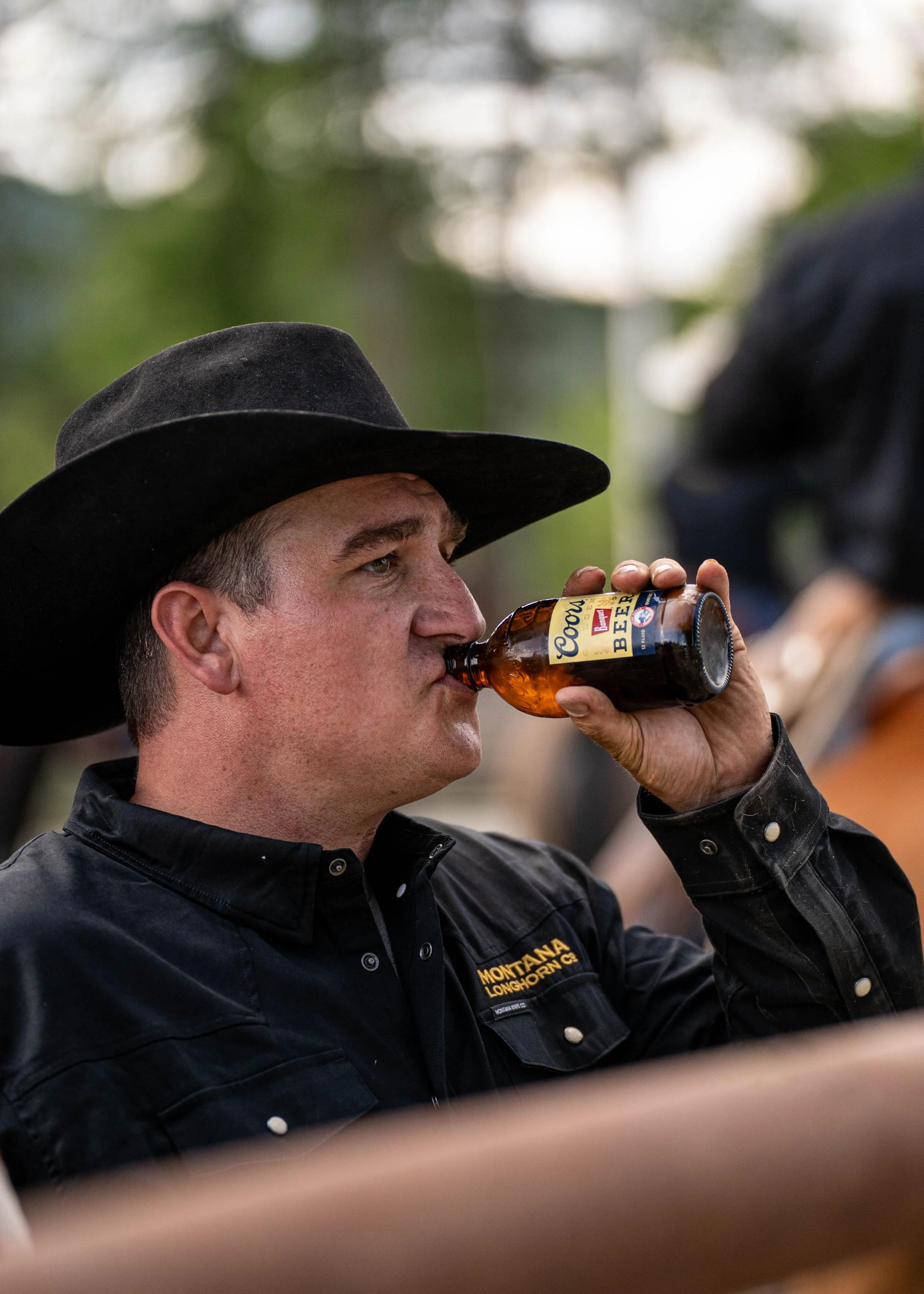 Cowboy wearing a black shirt and hat drinking a bottle of Coors beer at Montana Longhorn Ranch, captured during a ranch lifestyle photoshoot.