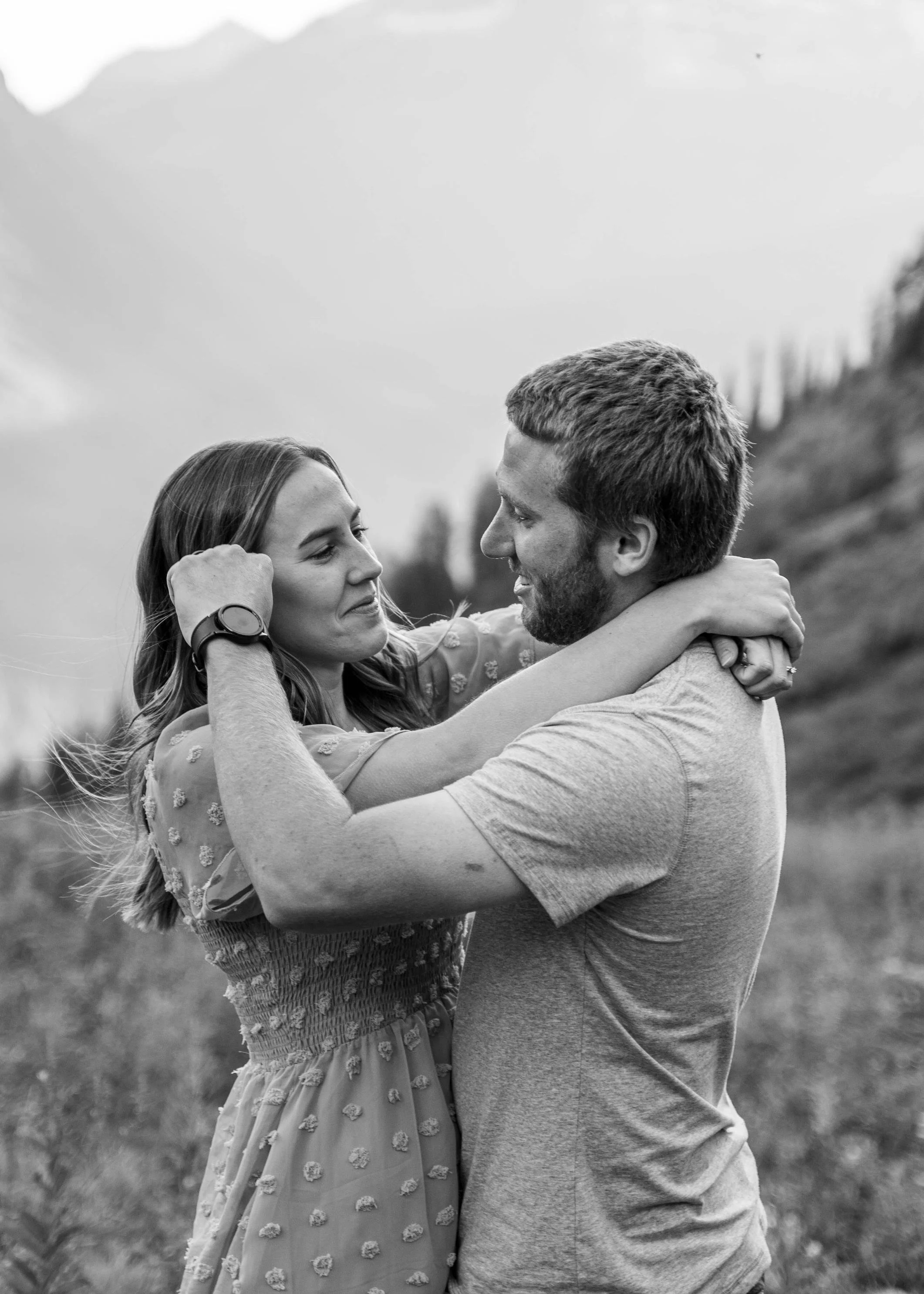 Black and white photo of a couple in a tender embrace, staring into each other’s eyes, with him gently moving her hair from her face, captured in Glacier National Park, Montana