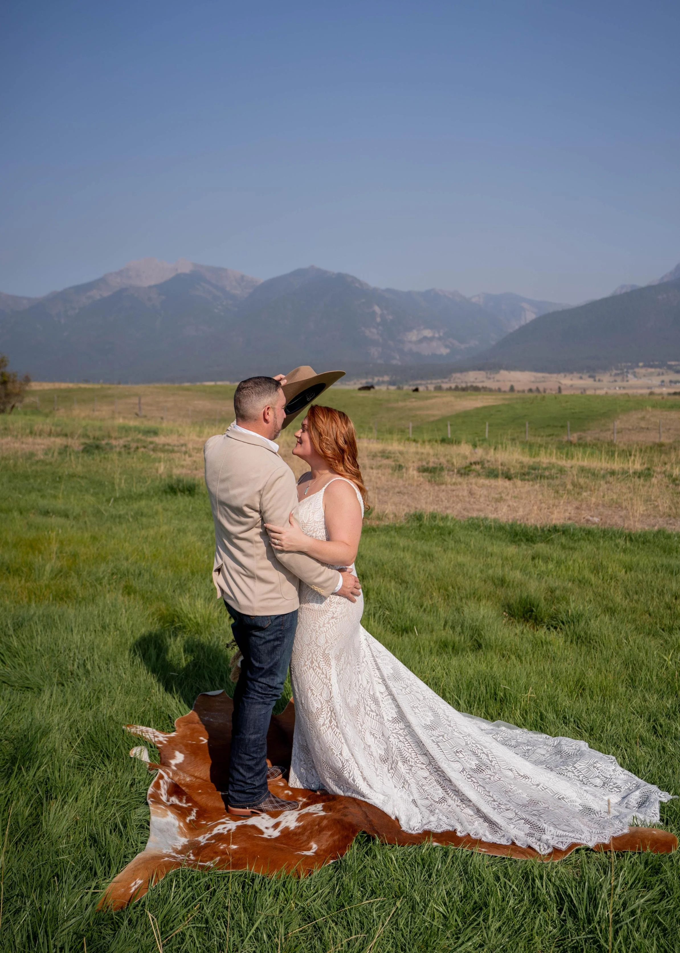 Groom holding his bride close and about to place his cowboy hat on her as they stand on a cowhide in a field, captured during a wedding photoshoot in Flathead Valley, Montana