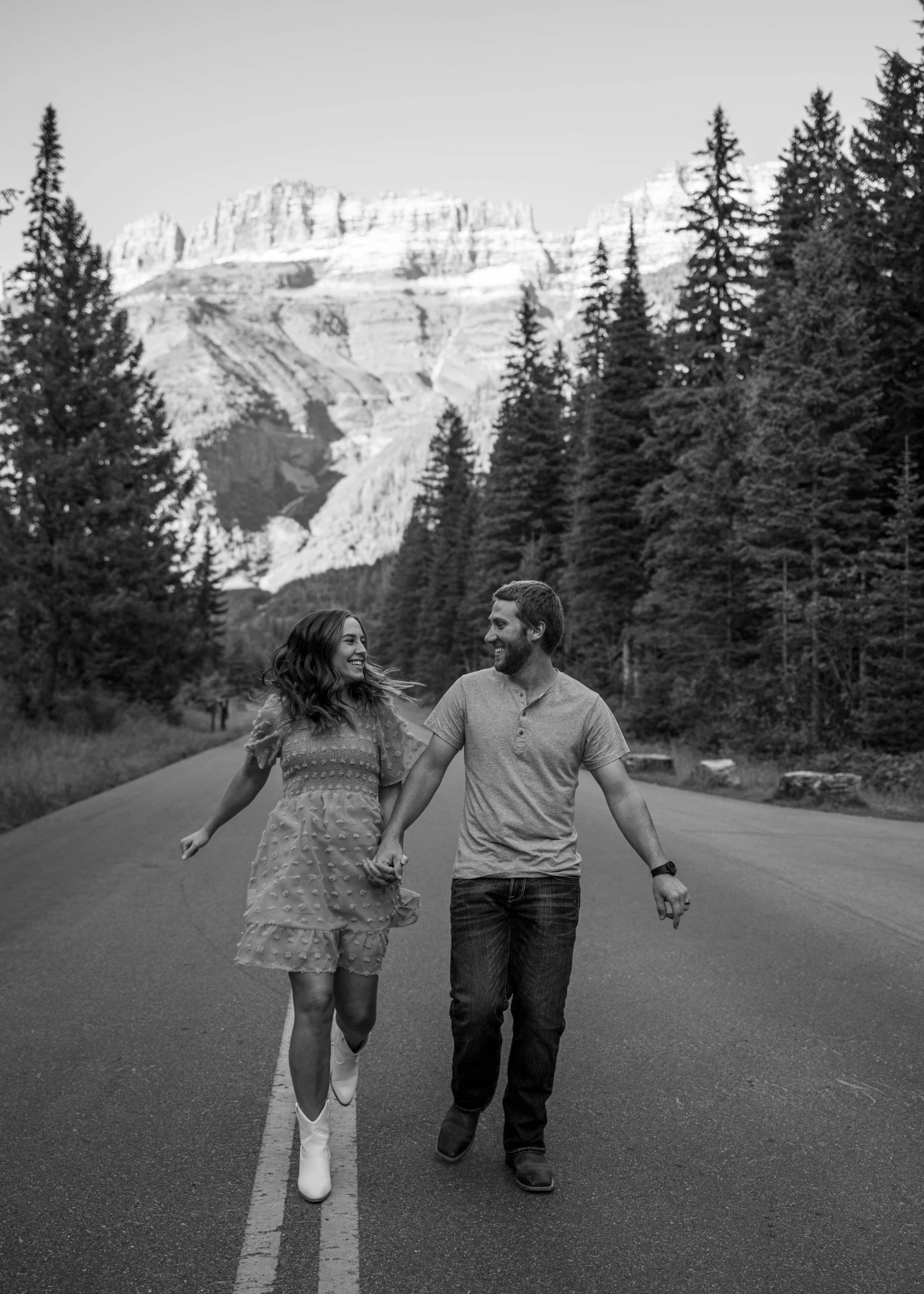 Black-and-white photo of a couple running hand in hand on a road in Glacier National Park, Montana, captured during a couples photoshoot