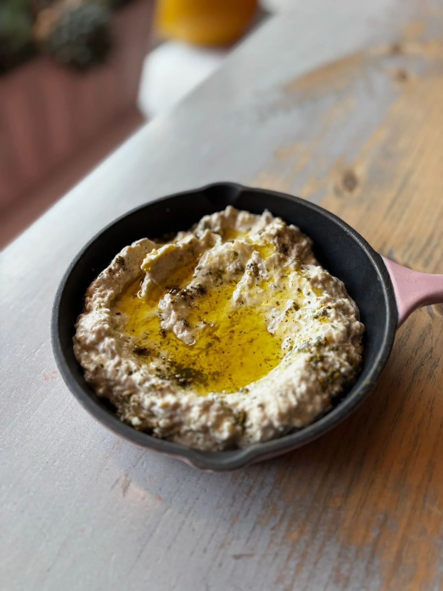 A small black skillet containing a creamy, cheese-topped dish with olive oil drizzled on top, placed on a wooden surface.