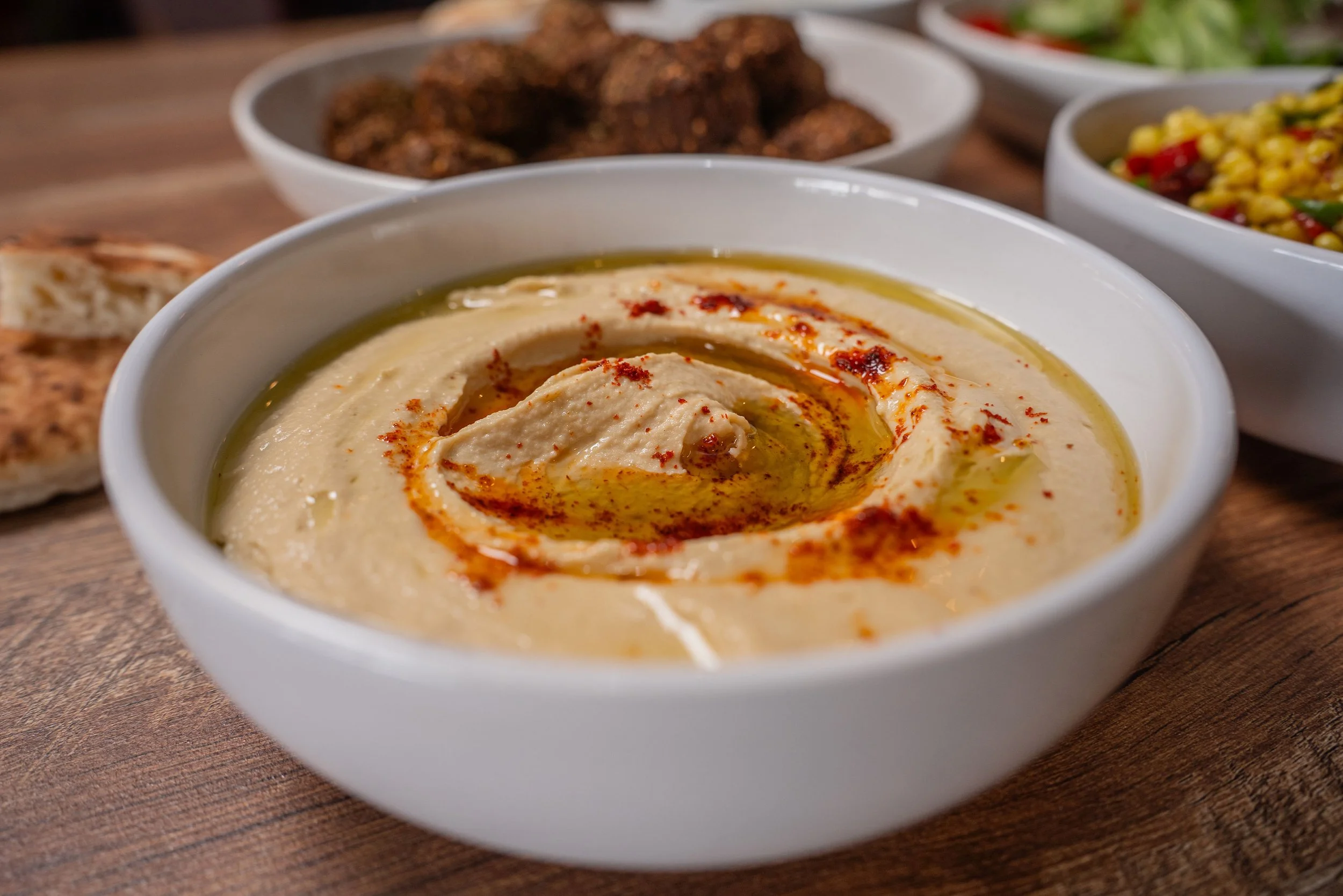 A bowl of creamy hummus garnished with paprika and olive oil, with other bowls of food in the background.