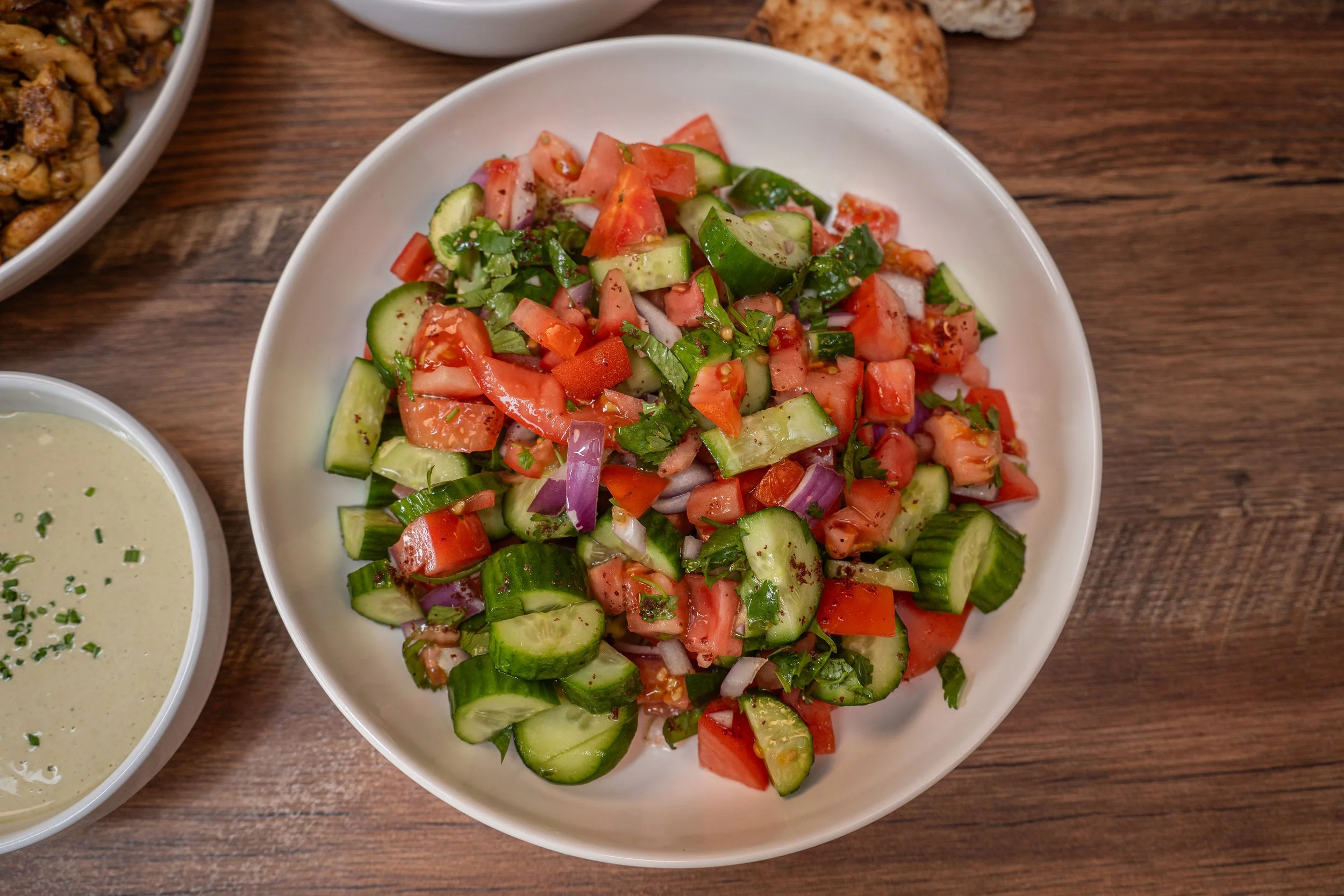 A white bowl filled with chopped fresh tomato, cucumber, onion, and herbs, likely a salad, on a wooden table. Part of a small bowl of salad dressing and another dish are visible around it.