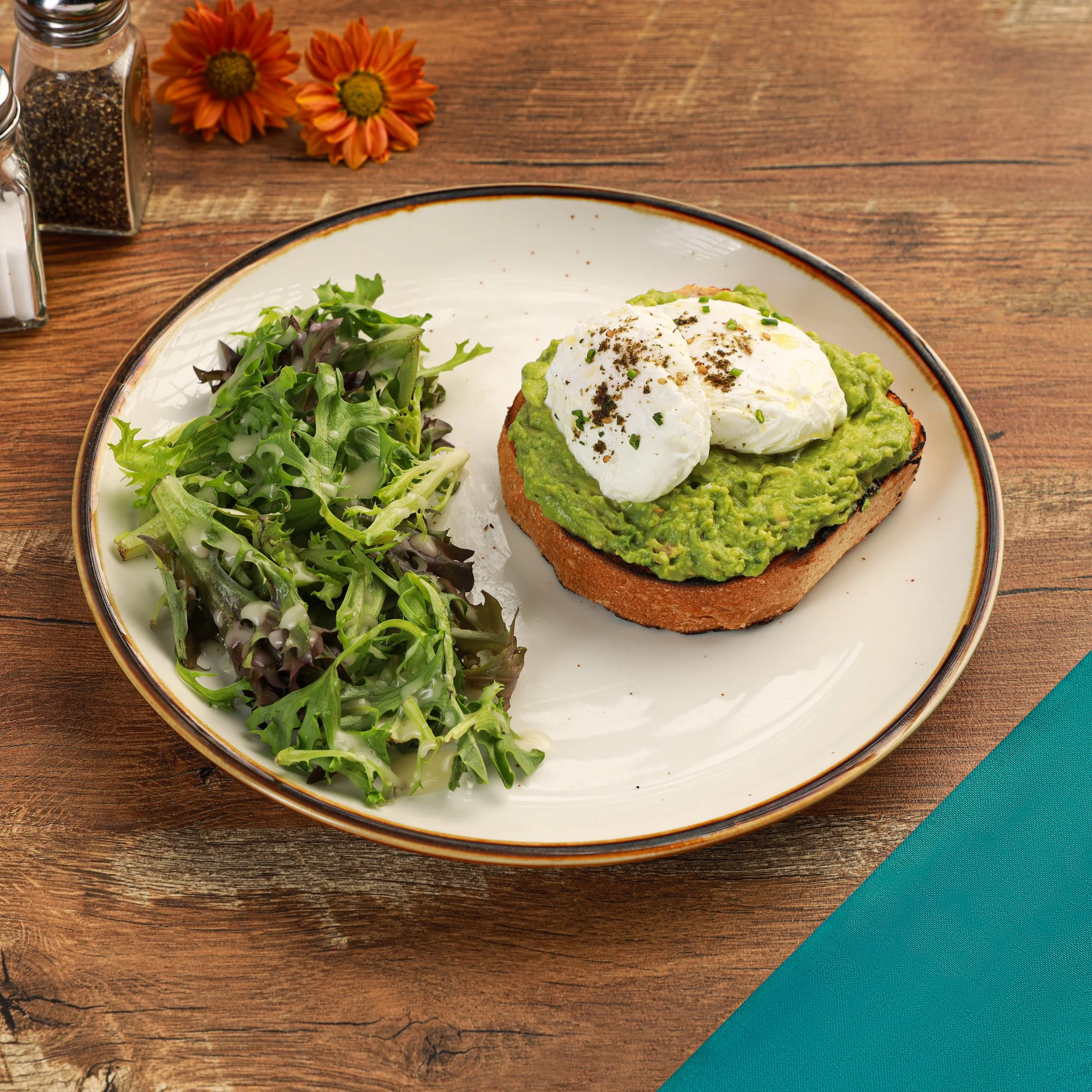 Dish of toast topped with mashed avocado, poached egg, and black pepper, served with mixed greens on a white plate. The plate is on a wooden table with a jar of pepper and flowers nearby.