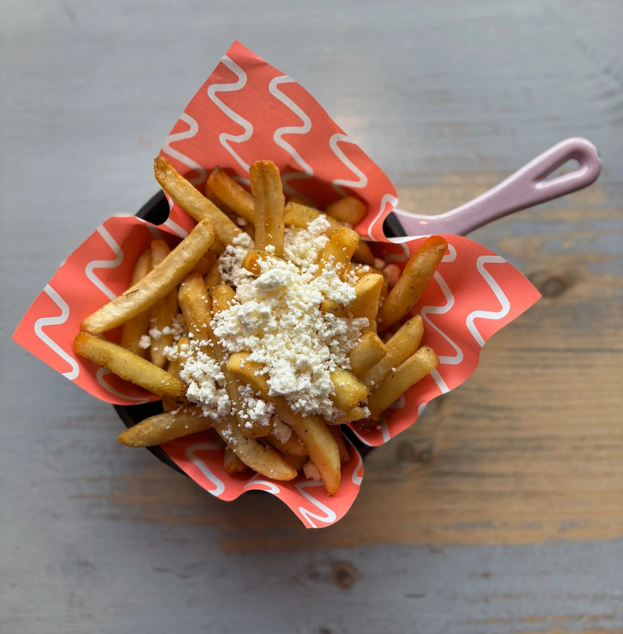 French fries inside red and white zigzag patterned paper in a black container, with a pink plastic fork resting on a gray wooden table.