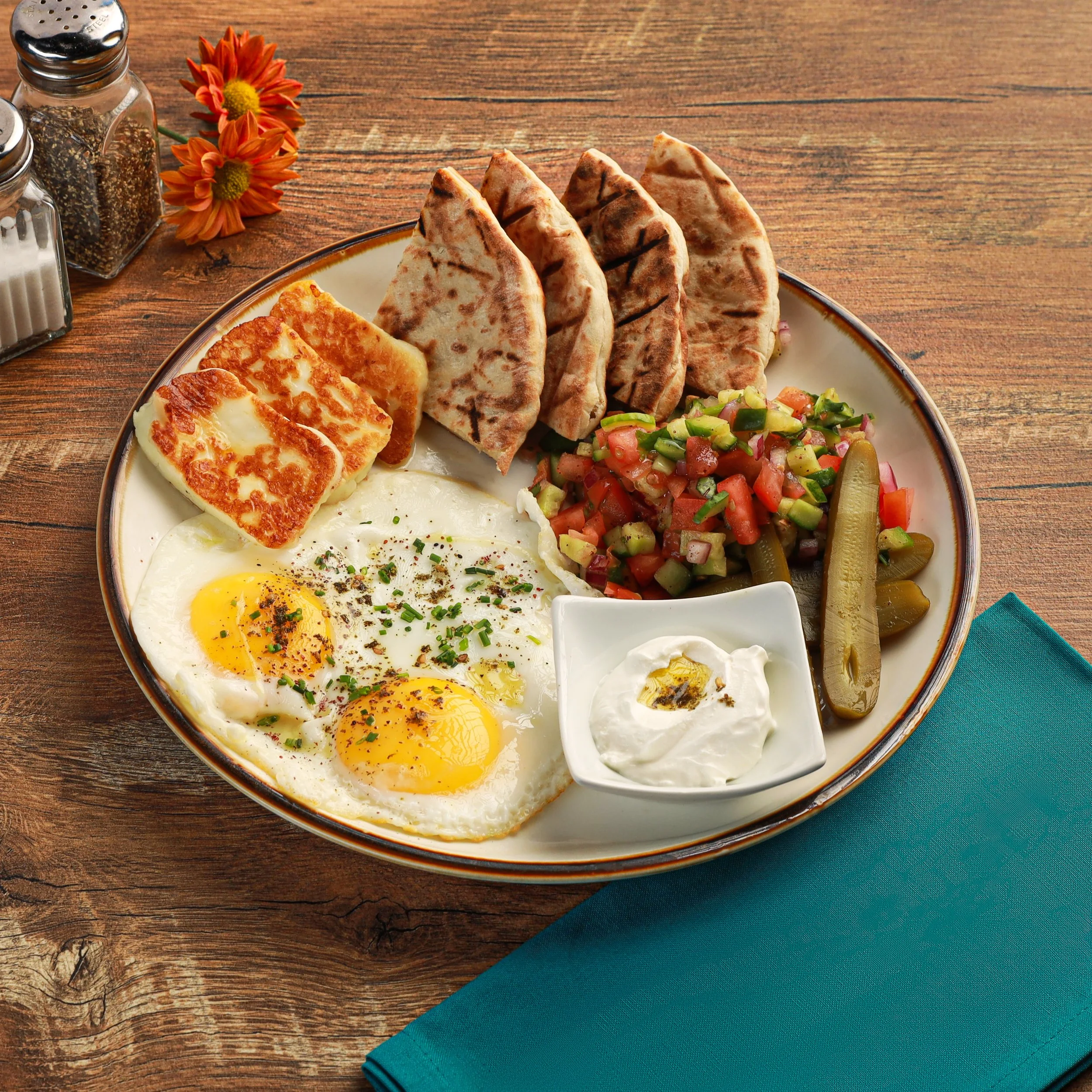 A white plate with sunny side up eggs, toasted flatbread, chopped vegetables, and pickles on a wooden table, with salt, pepper, and flower decorations nearby.