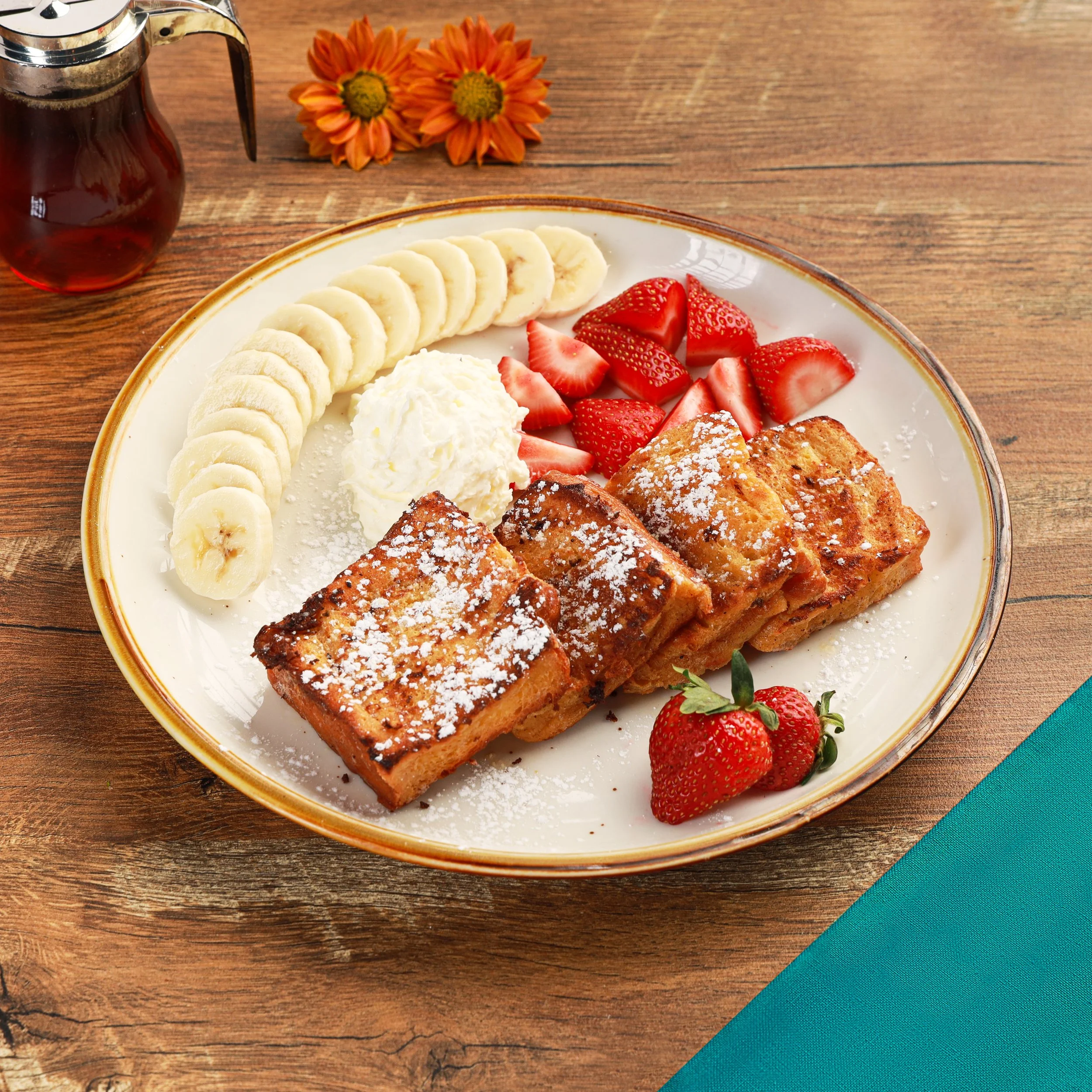 A plate of sliced bananas, strawberries, and fried French toast with powdered sugar, garnished with whipped cream on a wooden table. A jar of syrup and orange flowers are in the background.