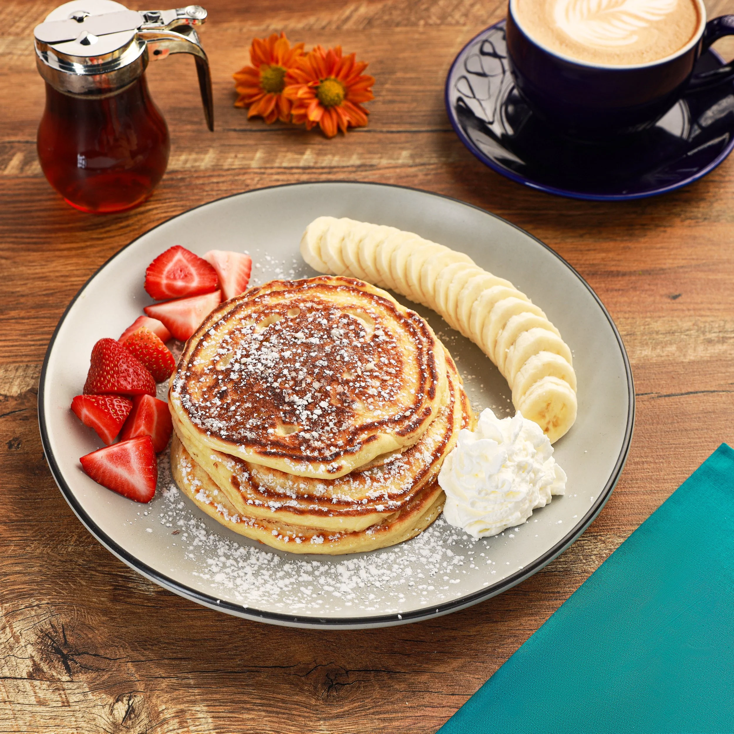Plate of pancakes topped with powdered sugar, sliced bananas, and strawberries, with a cup of coffee, honey jar, and orange flowers on a wooden table.