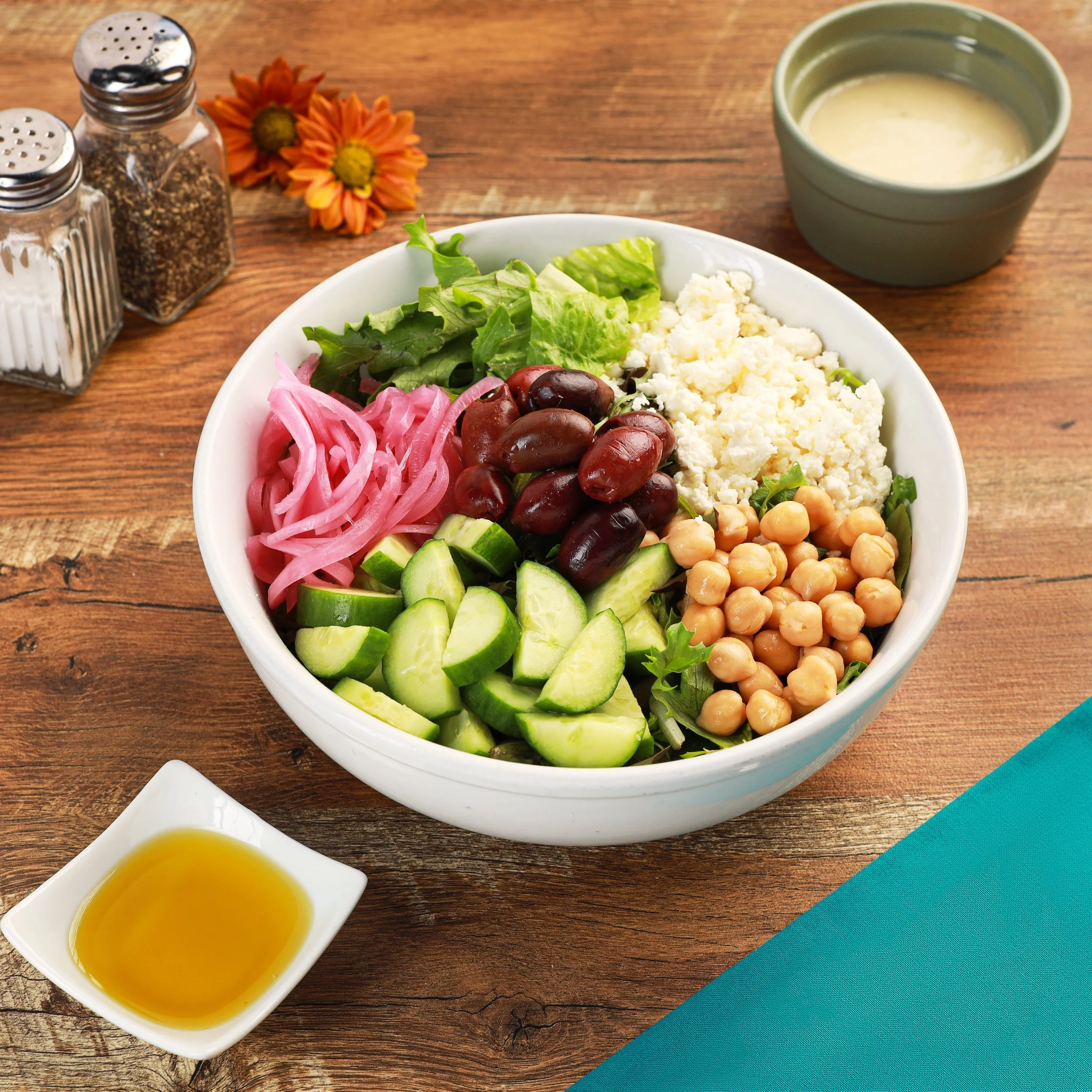 A bowl of fresh salad with lettuce, cherry tomatoes, pickled red onions, cucumber, feta cheese, and chickpeas on a wooden table, accompanied by a small dish of olive oil dressing, a pair of salt and pepper shakers, and two orange flowers.