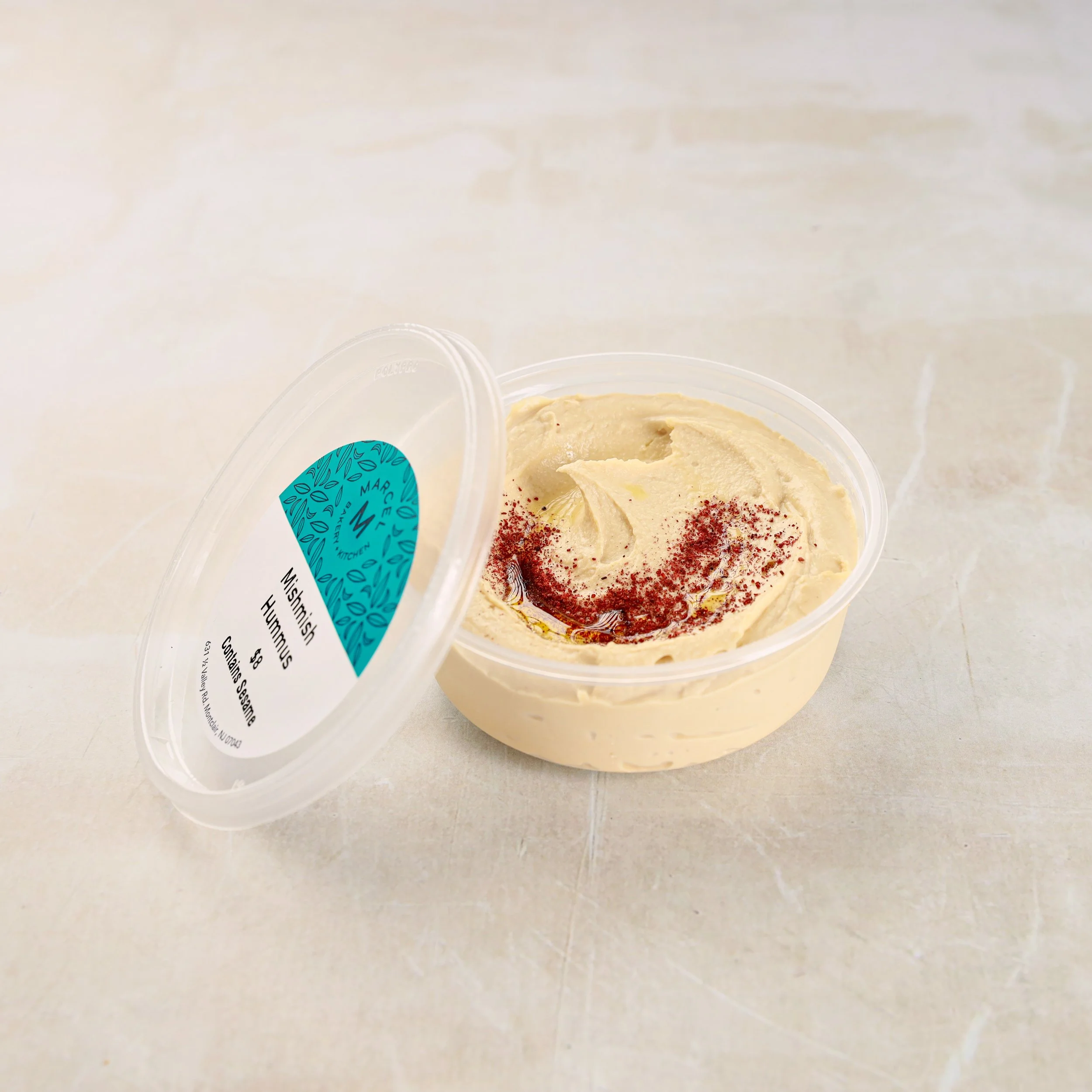 A clear container filled with a creamy, beige spread, garnished with red spice or seasoning, placed on a light-colored surface.