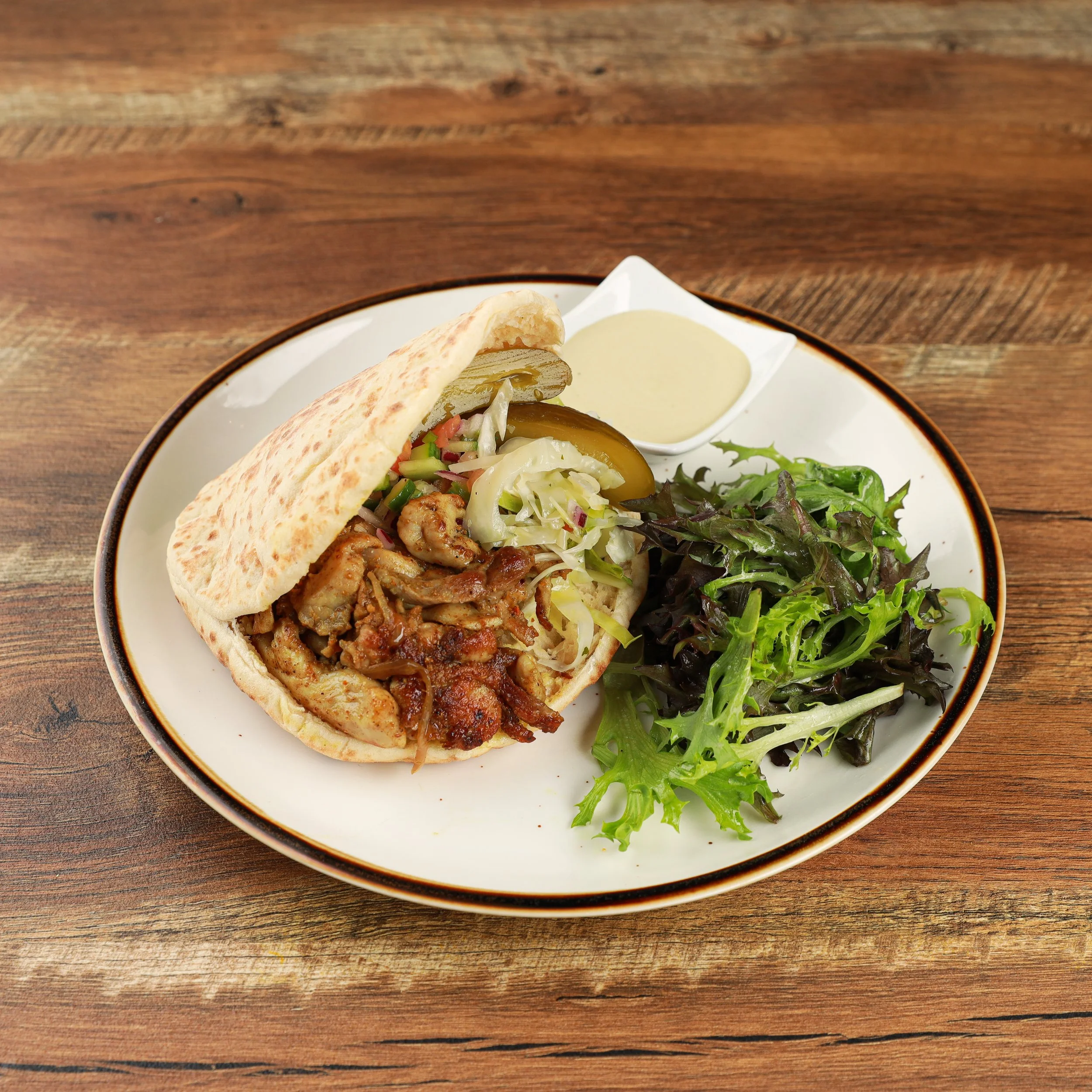 Close-up of a plate with grilled chicken Shawarma, fresh vegetable salad, and a side of sauce, placed on a wooden table.