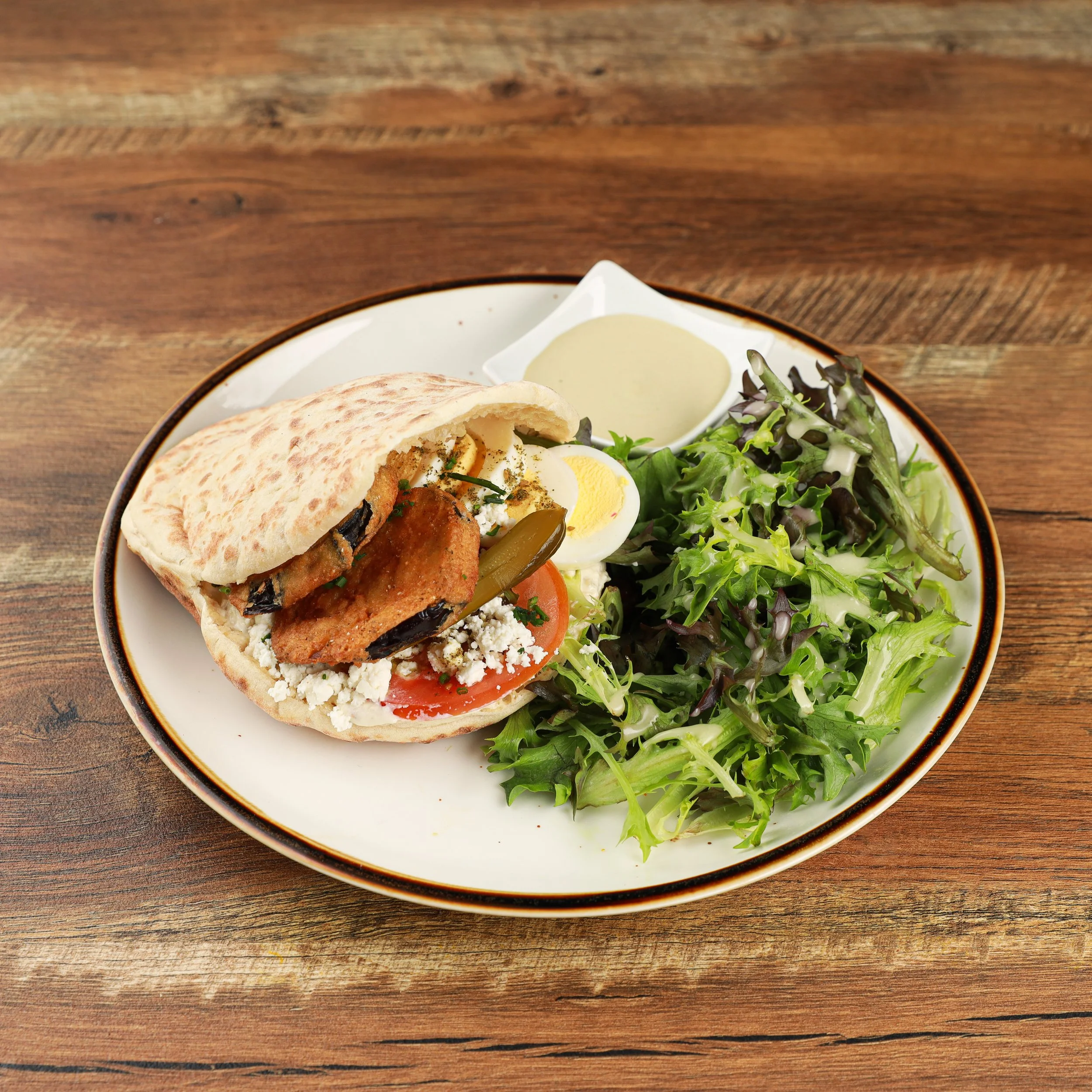Plate of food including pita bread, grilled chicken, boiled egg, tomato slices, pickles, and salad with mixed greens and dressing, on a wooden table.
