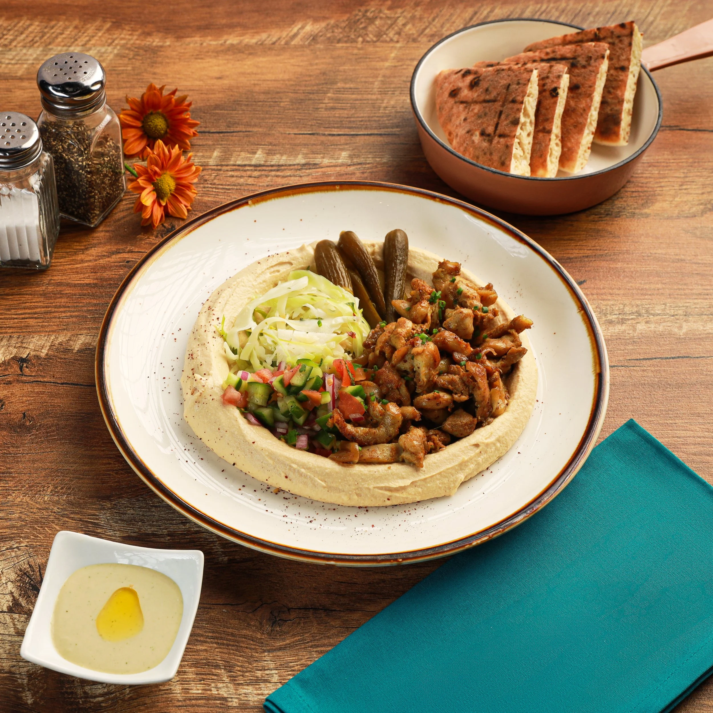 A plate of Middle Eastern hummus topped with chopped vegetables and a side of grilled pita bread with herbs, accompanied by a small dish of olive oil and lemon juice, on a wooden table with a teal cloth.