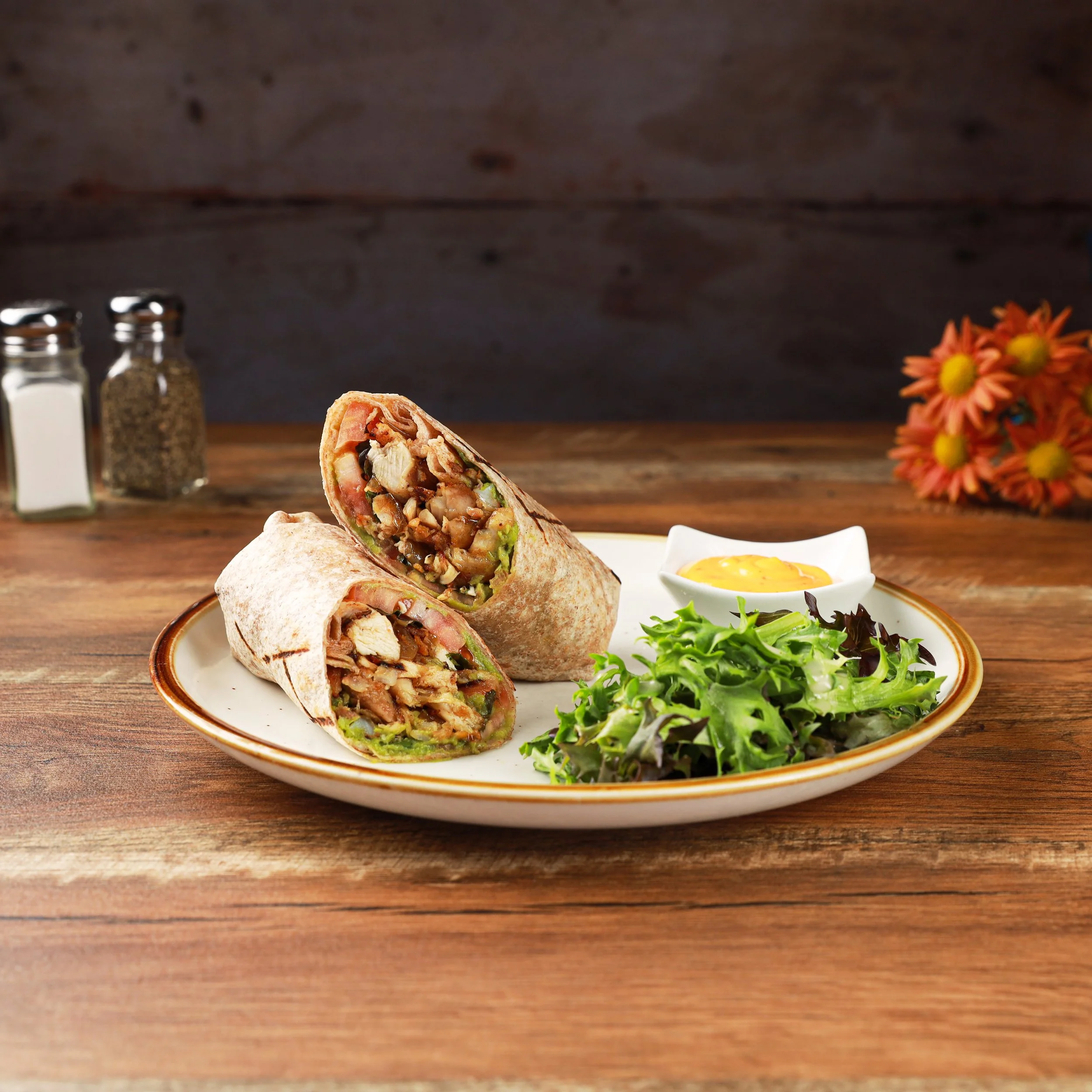 Close-up of a sliced wrap filled with chicken, vegetables, and sauce on a white plate, with a side salad, yellow dipping sauce, salt, pepper shakers, and a bouquet of orange flowers in the background on a wooden table.