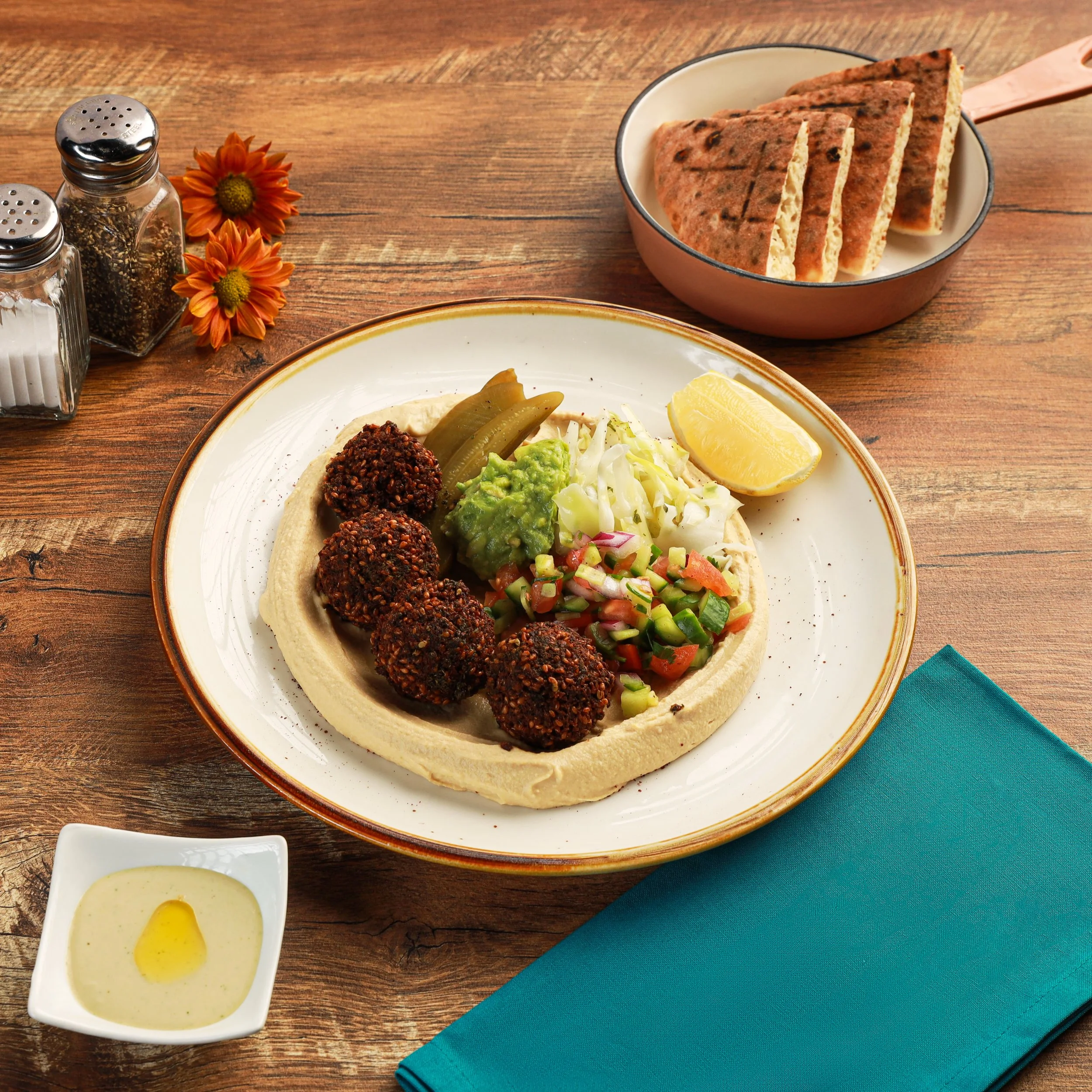 A plate of Mexican food with grilled peppers, guacamole, shredded cheese, and a lemon wedge; a small bowl of creamy dressing; a pink bowl with grilled flatbread; salt and pepper shakers; orange and yellow flowers; a blue napkin on a wooden table.