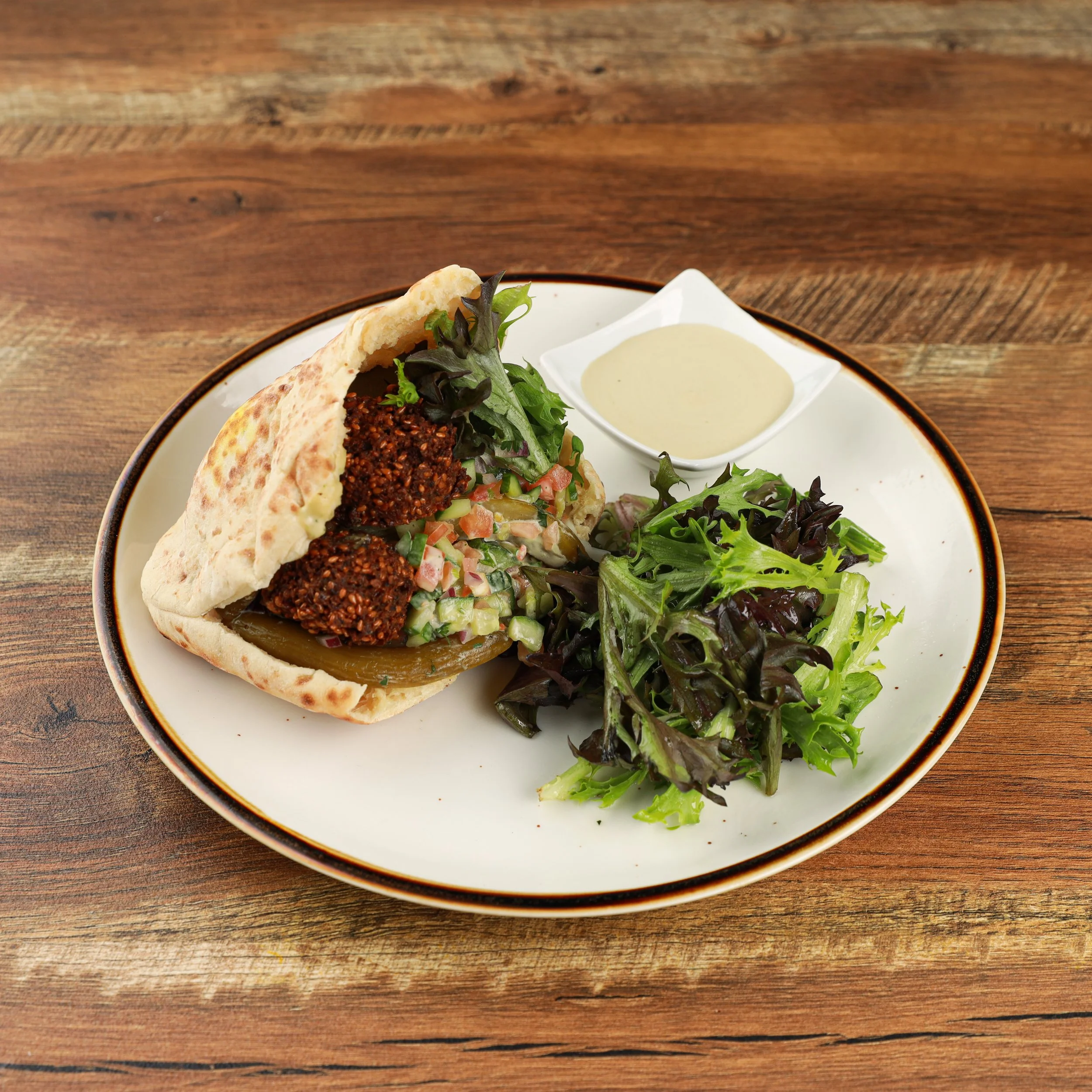 A plate with pita bread, tabbouleh salad, falafel, mixed greens, and a side of tahini sauce on a wooden table.