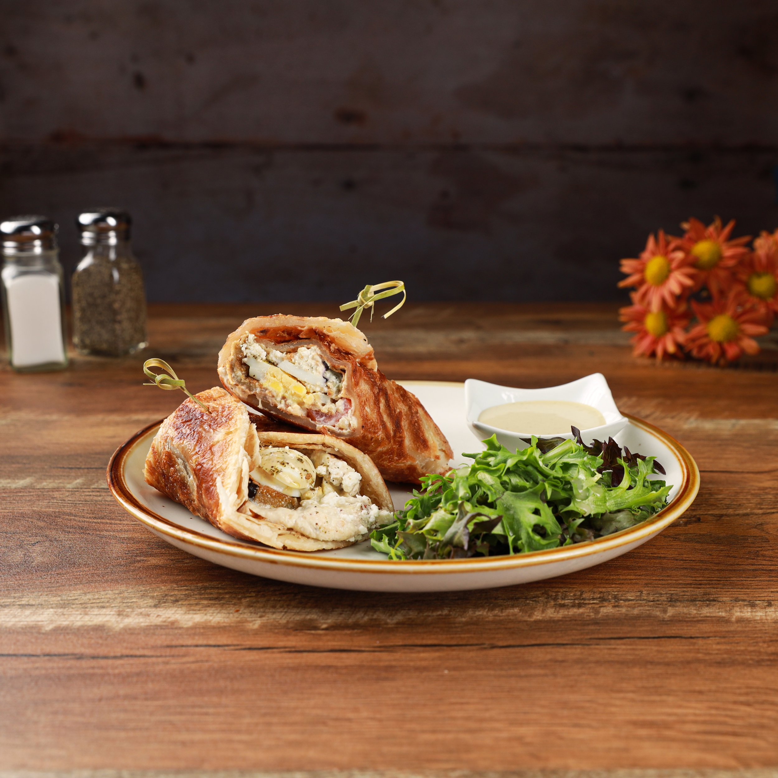 A plate of savory pie with a cut piece showing a filling of cheese and meats, accompanied by a small dish of sauce, on a wooden table with salt and pepper shakers and a flower arrangement in the background.
