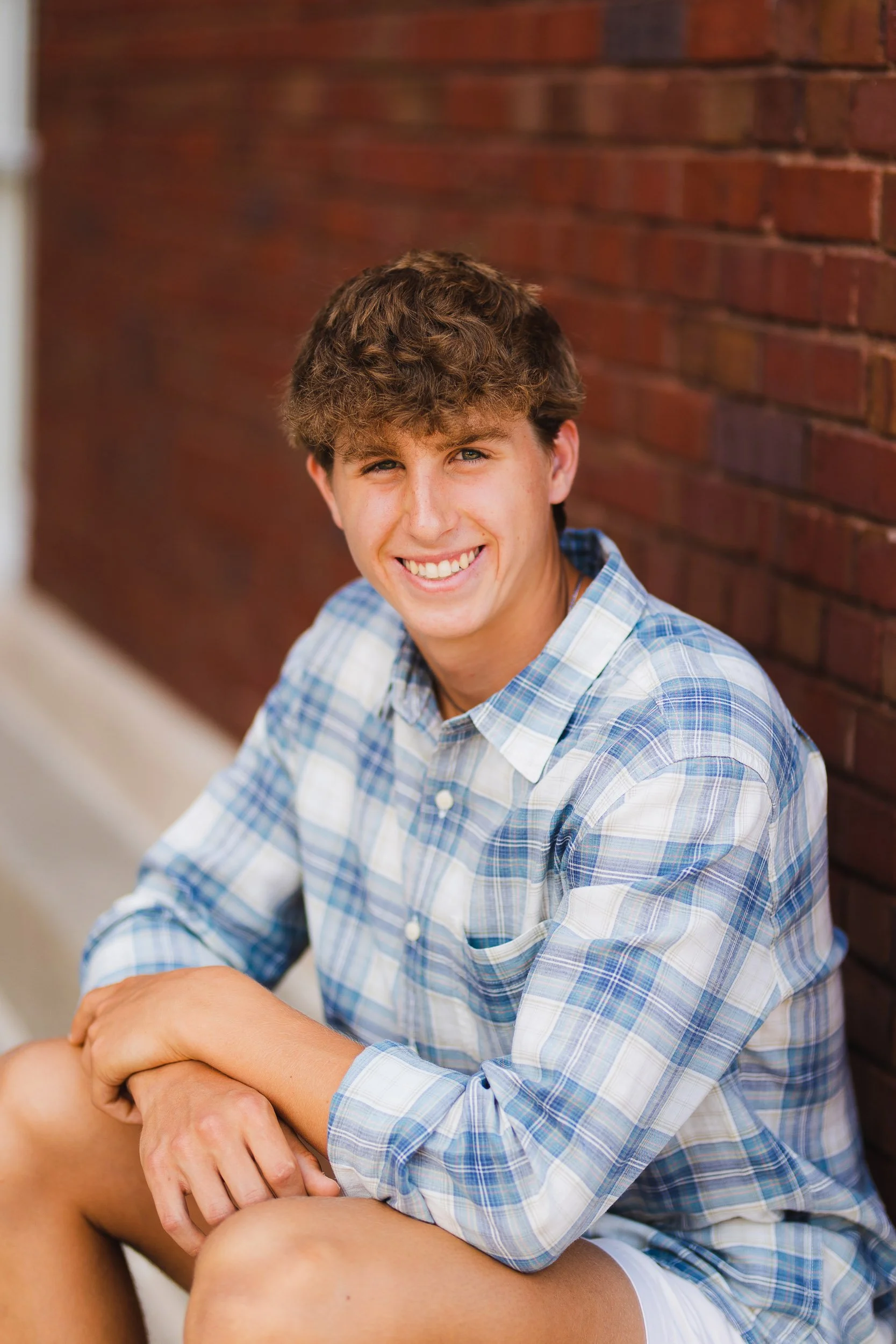 Senior boy sitting against brick wall in plaid shirt, smiling