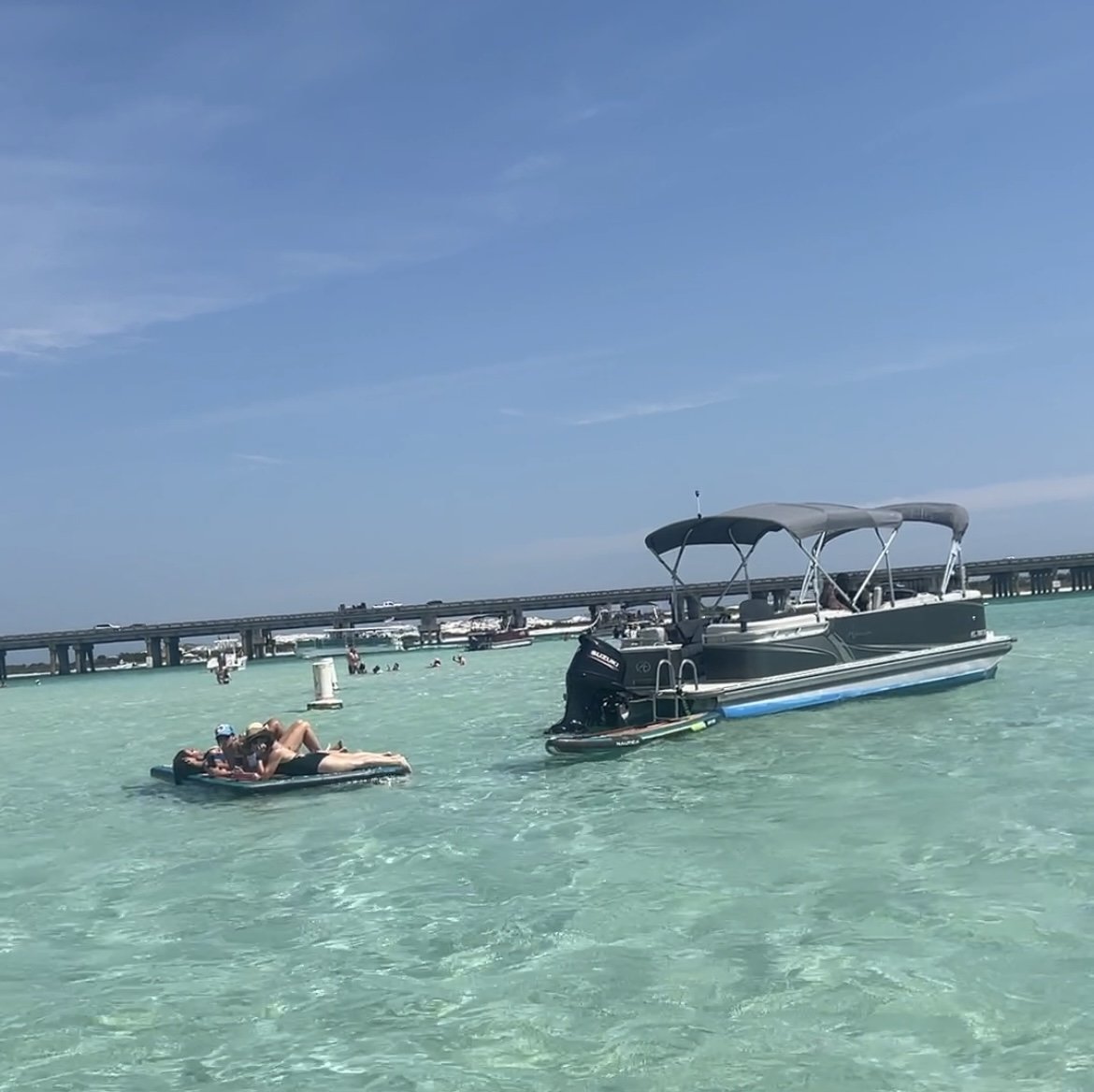 A boat and a person on a float in clear, turquoise water near a bridge on a sunny day.