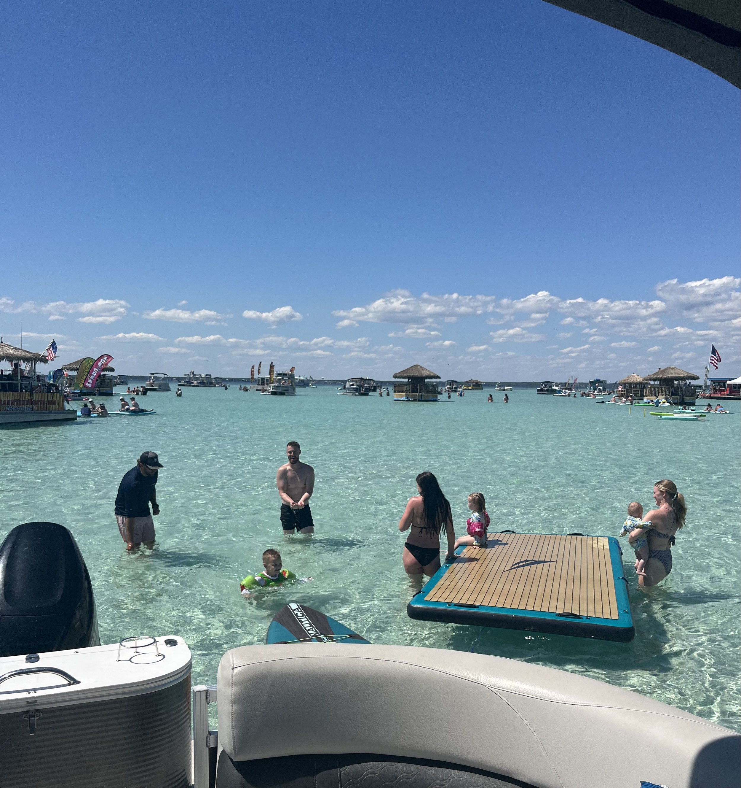 People enjoying a day on a clear, sunny beach with turquoise water, some wading and others on boats, and a floating dock in the water.