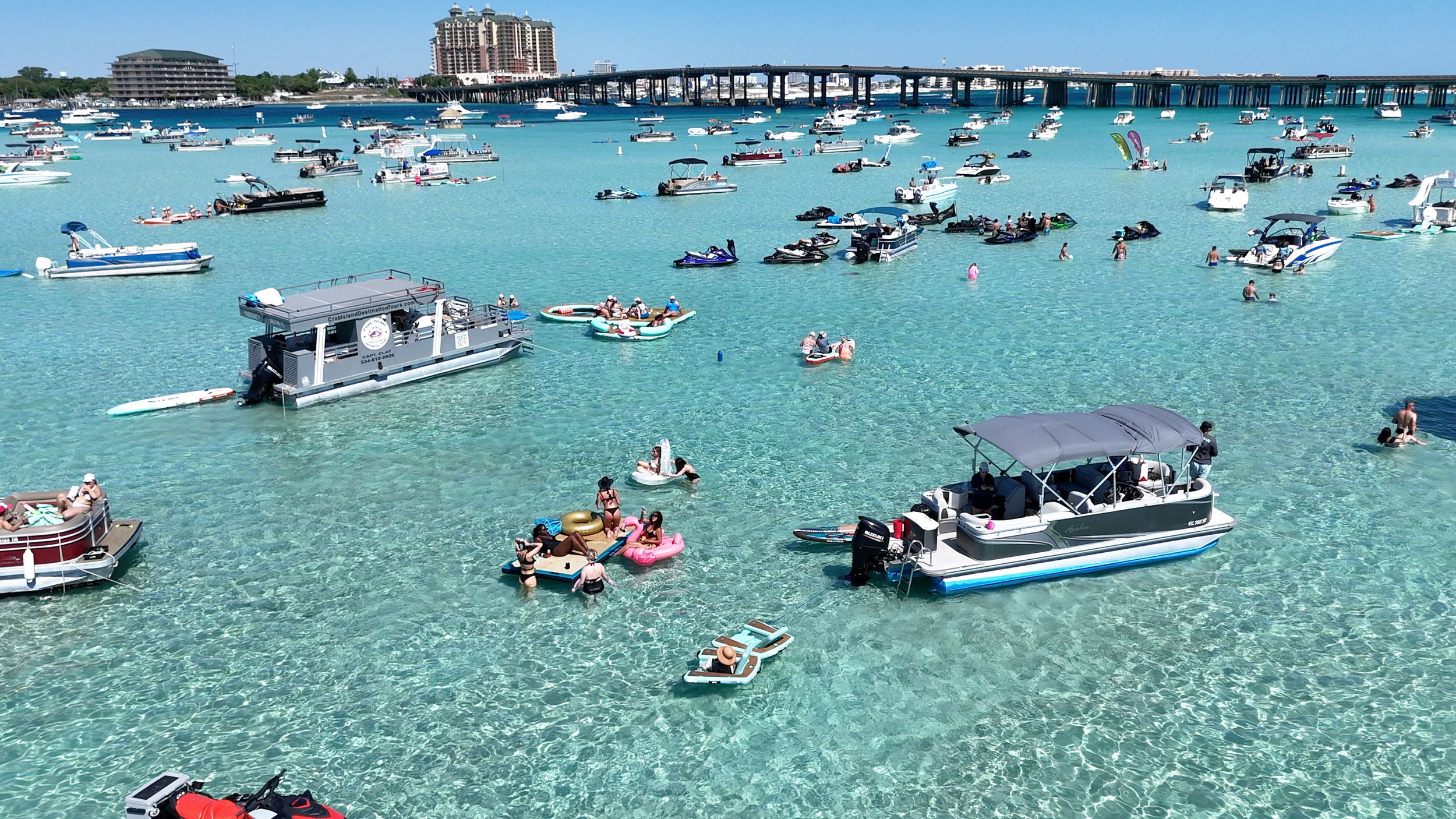People swimming and relaxing on inflatable floats in clear turquoise water, with boats anchored nearby and a cityscape and bridge in the background.