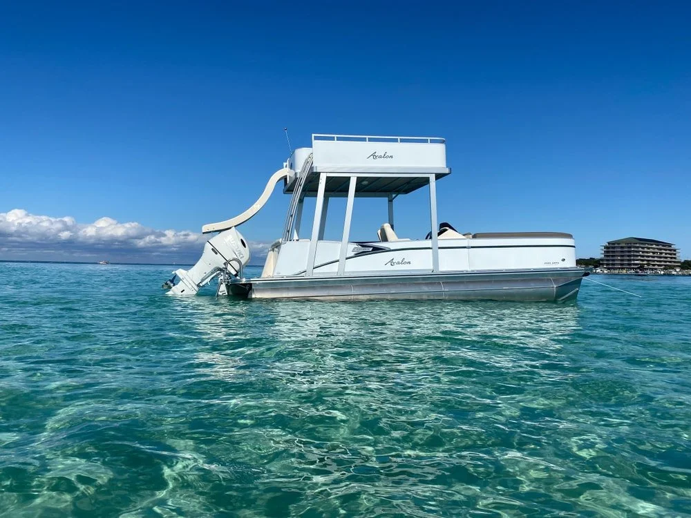 A white pontoon boat with an outboard motor partially submerged in the water, floating on clear turquoise ocean water under a blue sky with some clouds in the distance.