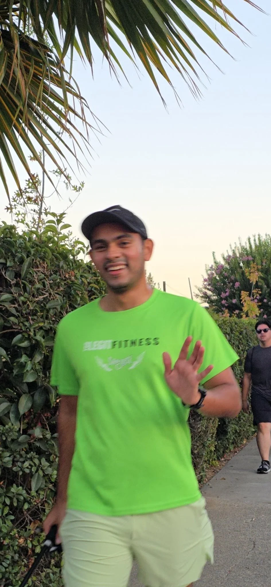 A smiling man in a neon green T-shirt and a black cap waving at the camera, walking on a sidewalk with bushes and trees in the background, and another person walking behind him.