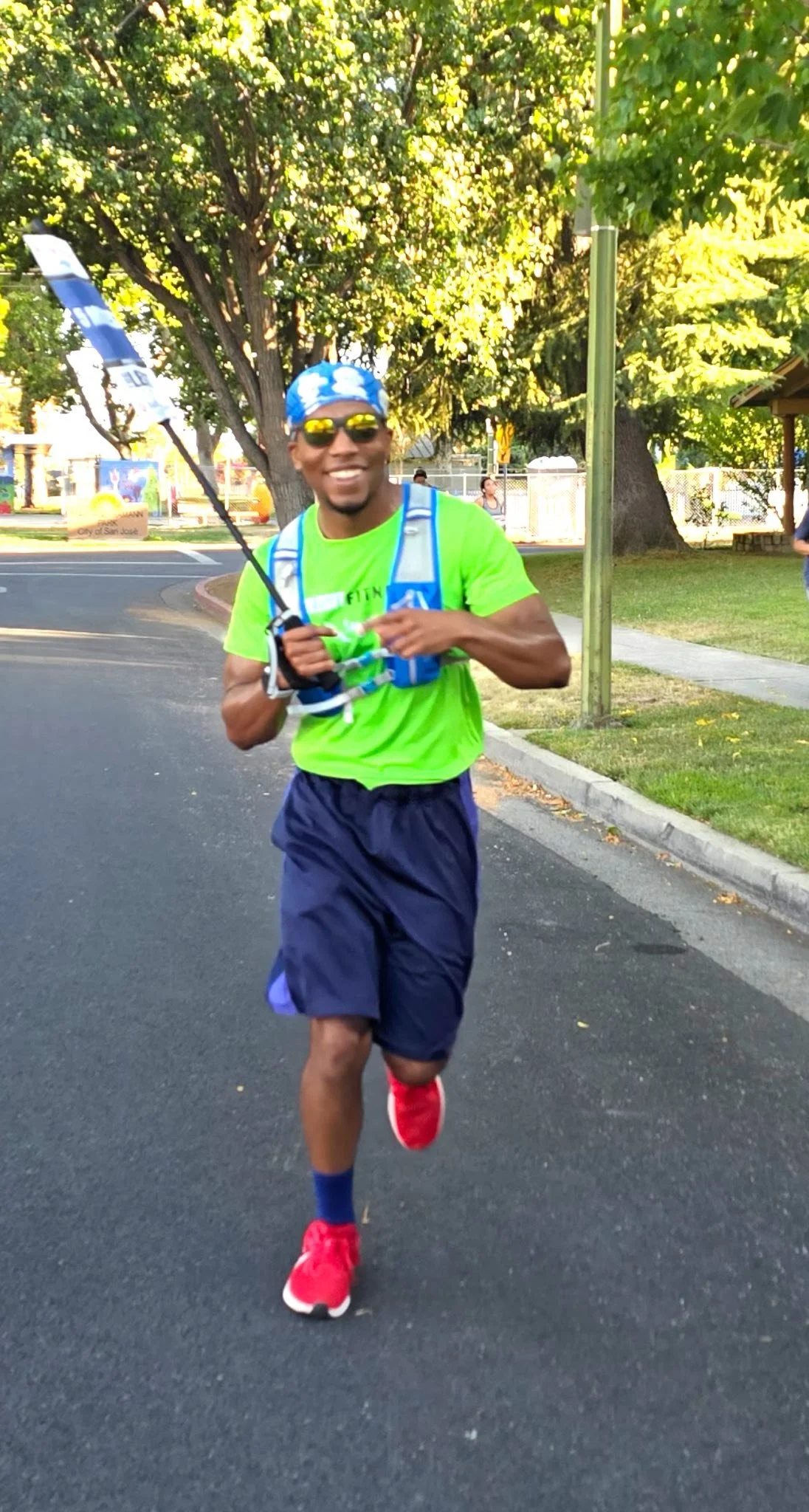 A man running outdoors during daylight, wearing a blue bandana, sunglasses, a neon green shirt, navy shorts, red running shoes, and carrying a blue hydration pack. He's holding a small flag and smiling.
