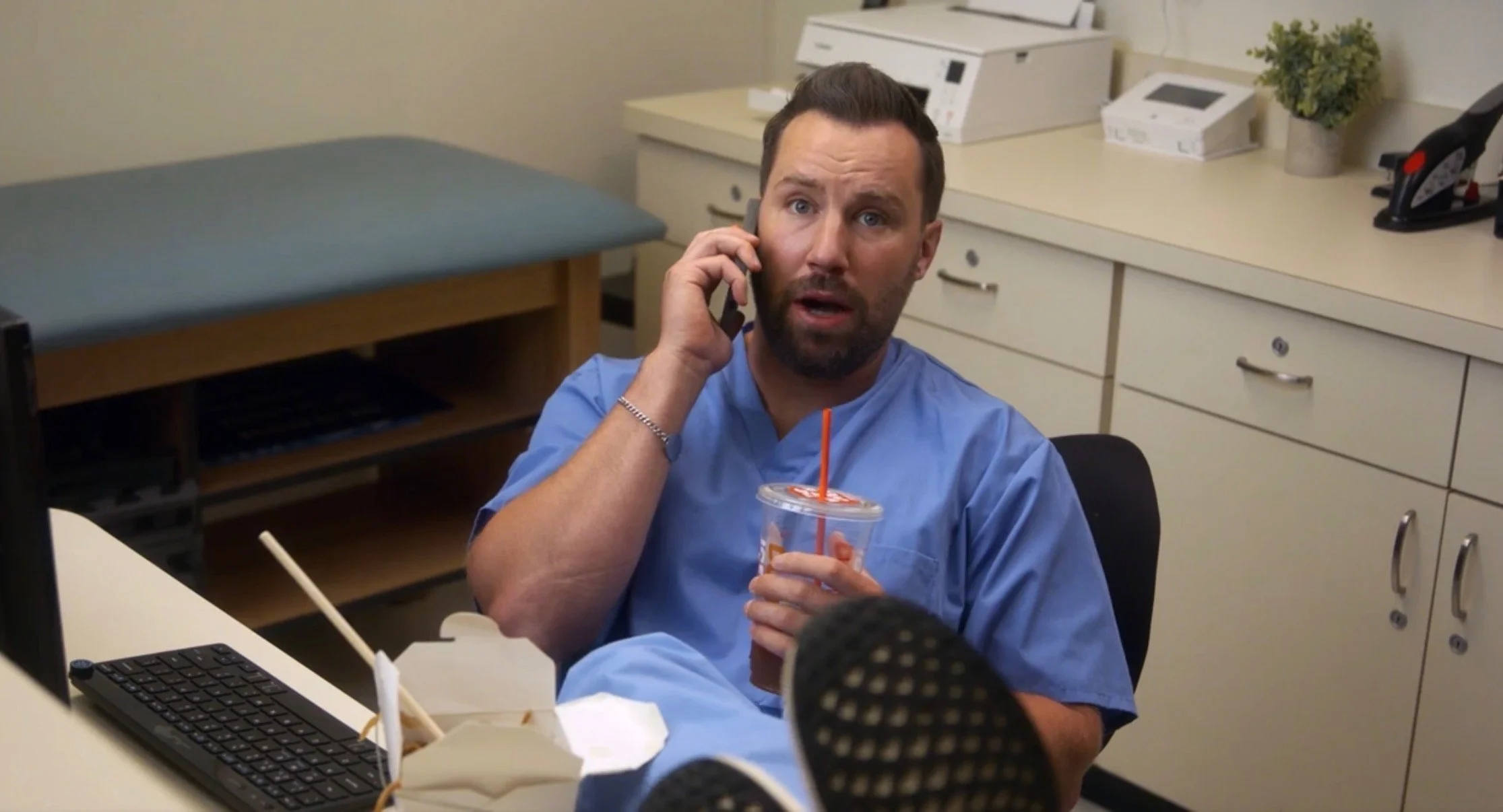 A man in light blue scrubs is sitting at his desk in an office, holding a cup with a straw in one hand and talking on a phone with the other hand. His feet are resting on the desk, showing the bottom of his shoes. The background includes office supplies, a computer, and a small potted plant.