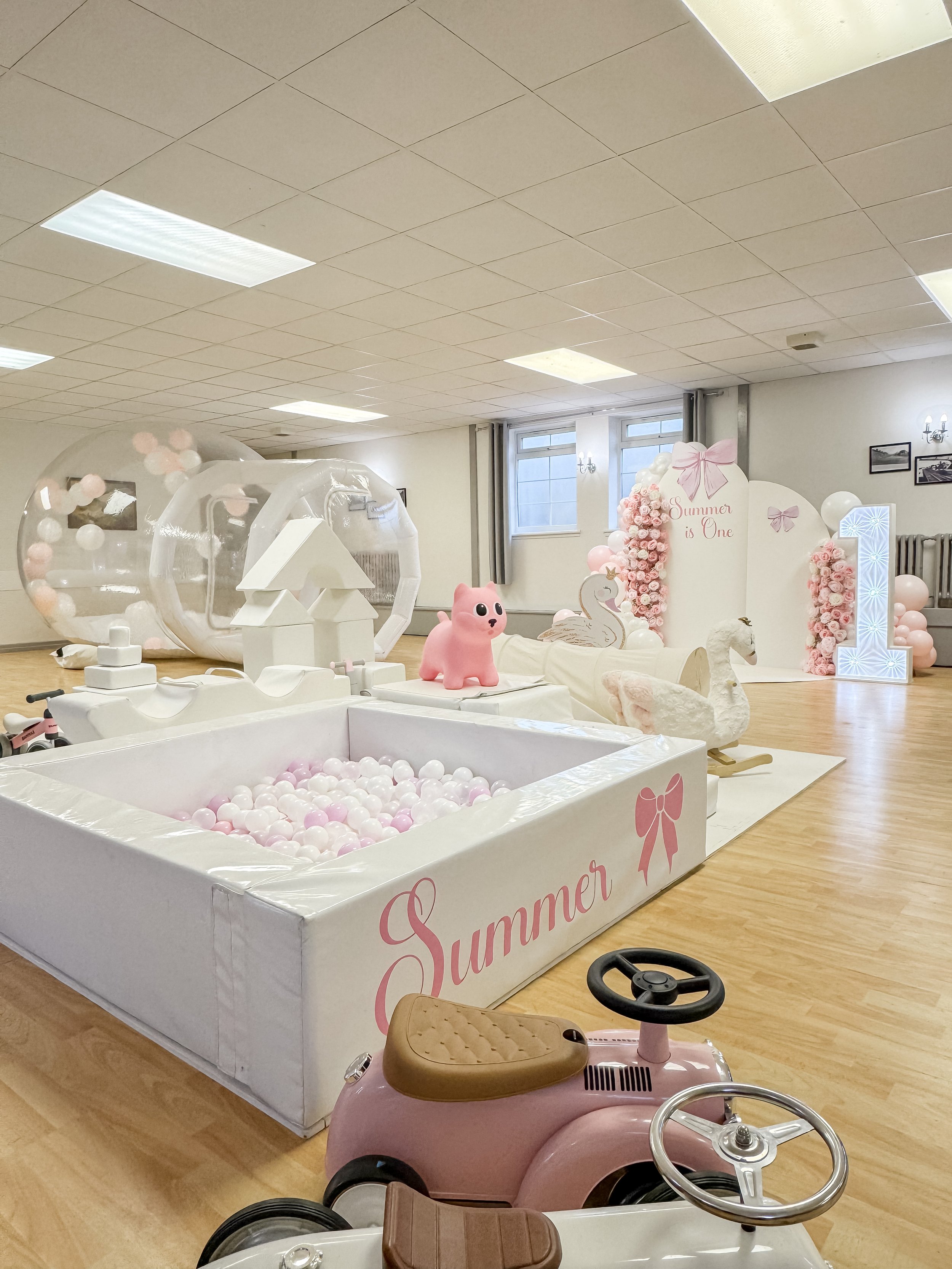 Decorative indoor setup with pink and white themed balloons, toys including a pink ride-on car, inflatable bubble tunnels, swan figures, and a large illuminated number one, all with the word "Summer" and pink bows, likely for a child's birthday celeb