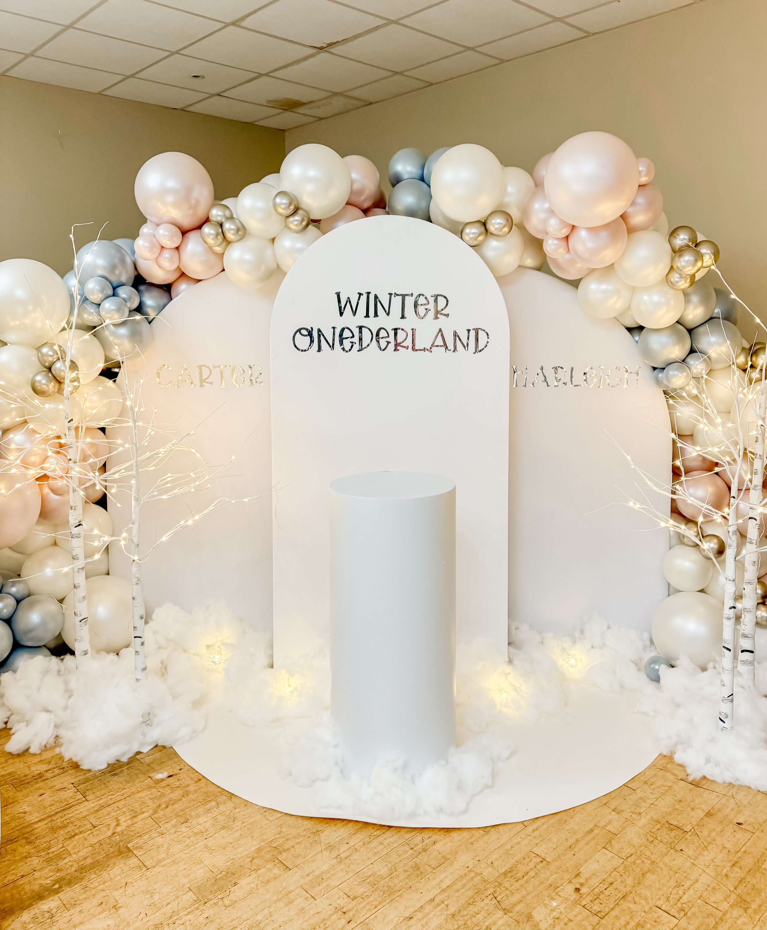 Winter Wonderland backdrop with balloons in white, silver, and pink, decorative white trees with string lights, and snow-like cotton base, featuring signs with the words 'Winter Wonderland', 'Carter', and 'Haleigh' for a festive event.