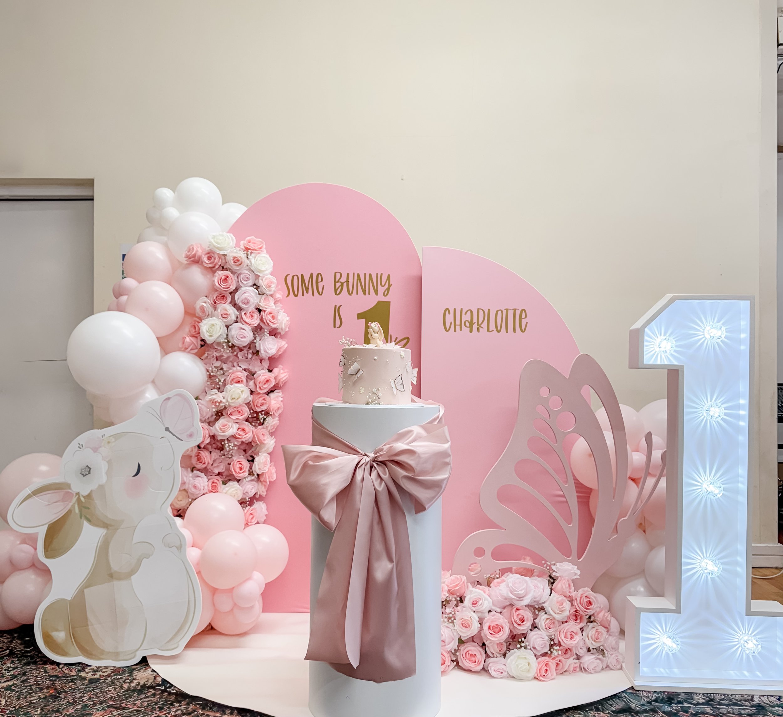 Pink and white birthday decorations for a girl, featuring balloons, flowers, a cake with a bunny topper, a large illuminated number one, and a bunny and butterfly cutouts.