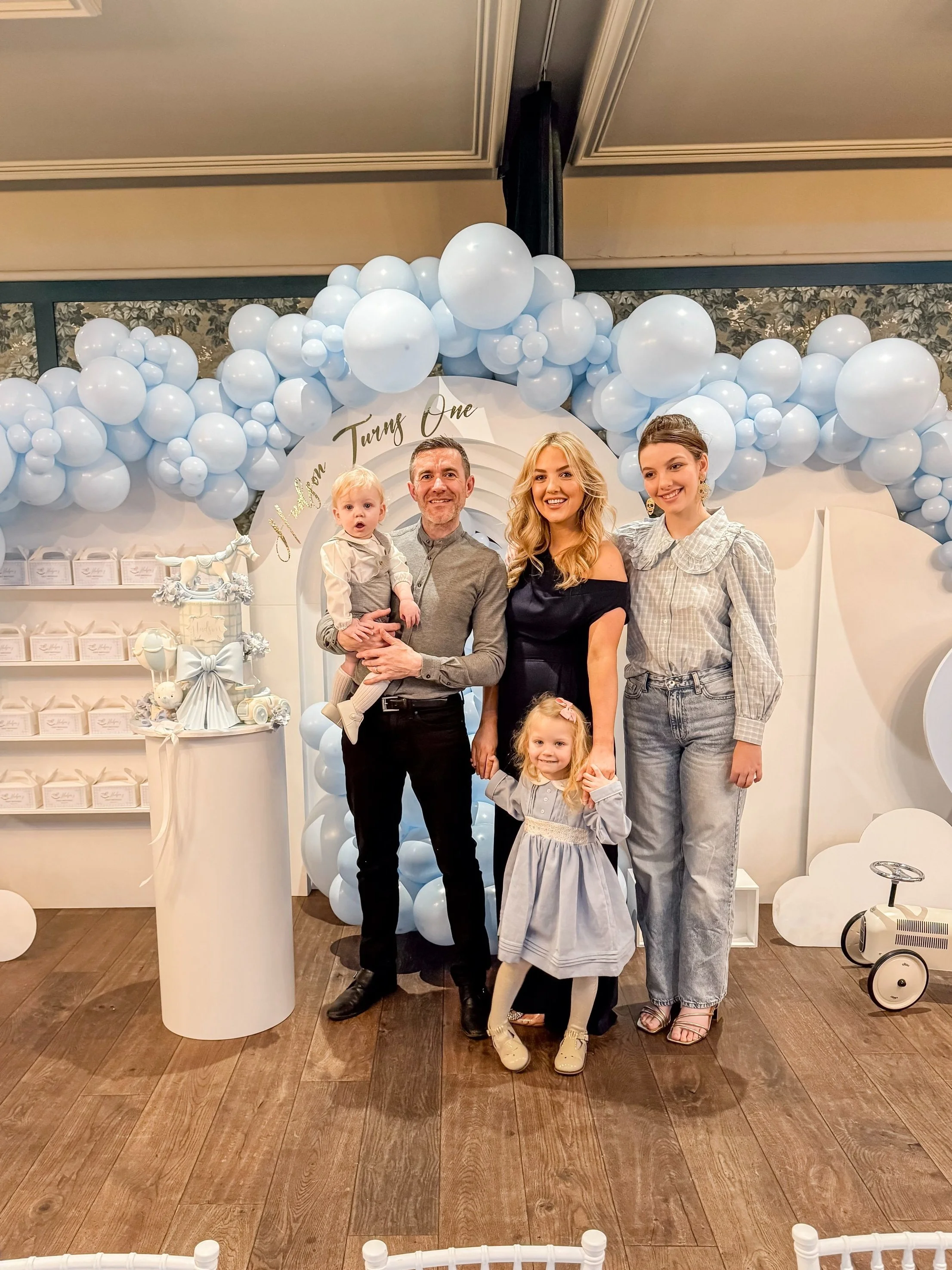 Group of five people, two adults and three children, standing together at a celebration with blue and white balloon decorations behind them. One child is being held by an adult. The background features a circular arch and a sign that says 'One.'