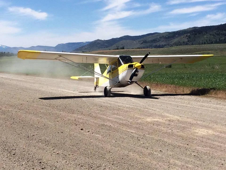 Small yellow and white propeller airplane on dirt runway with mountains and blue sky in background.