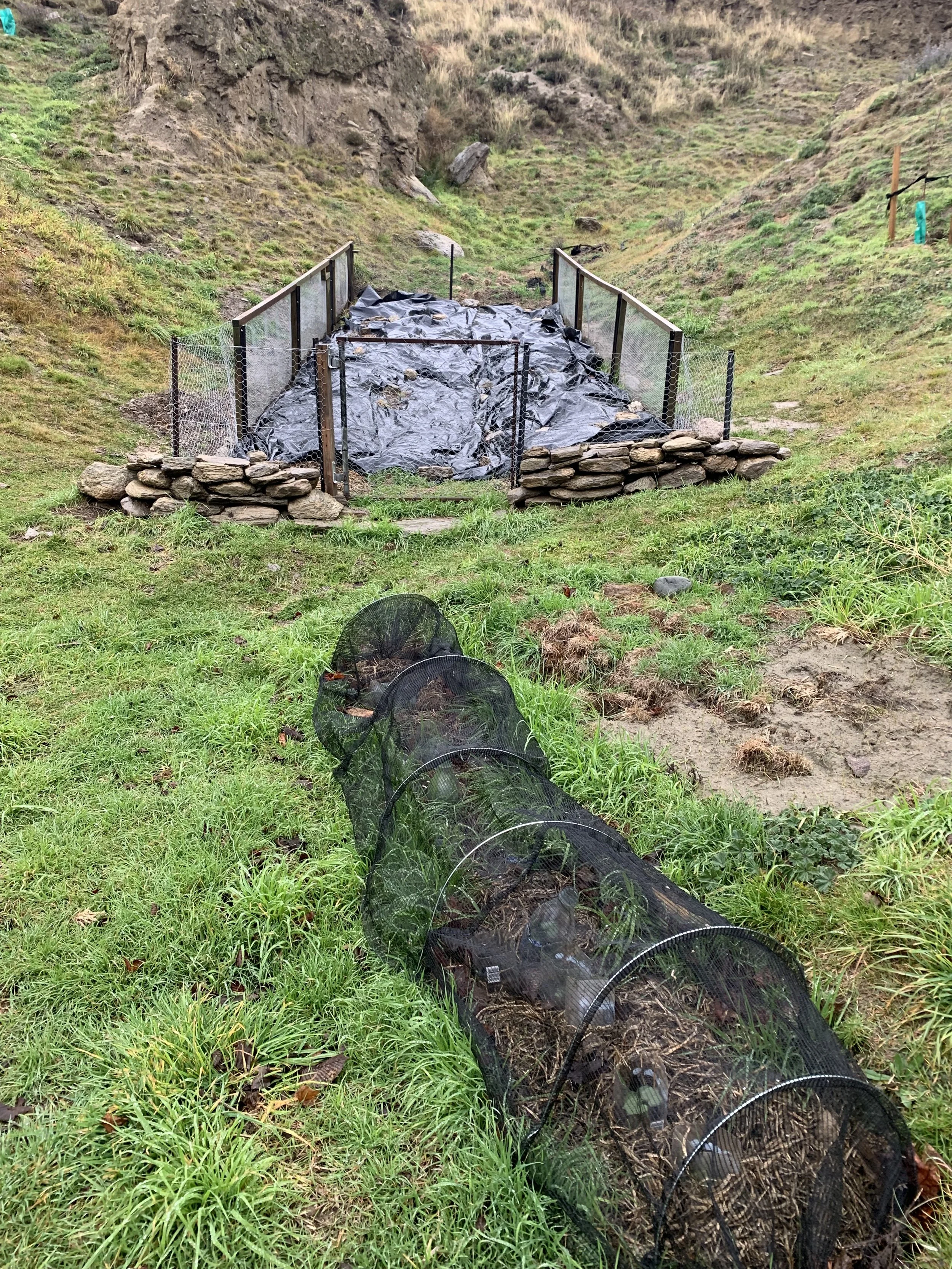 A small garden protected by rustic stone walls and a black metal fence, containing black plastic sheeting, situated in a grassy, hilly landscape with sparse vegetation.