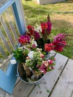 A metal bucket filled with a variety of colorful flowers, placed on a wooden porch near a blue railing, with green grass in the background.