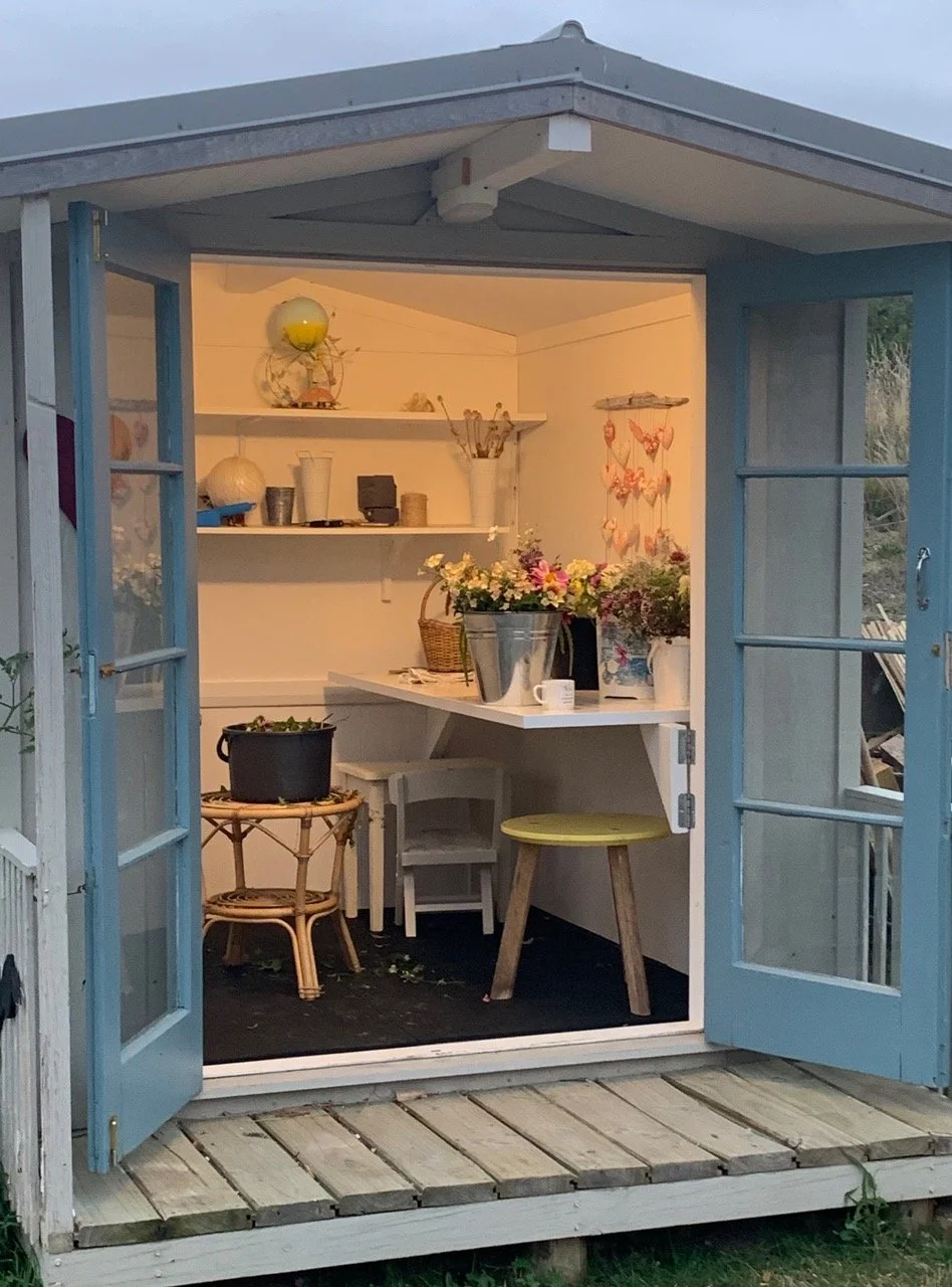 Small outdoor shed with open blue double doors, interior shelves with decorative items, vases, and flowers, small table with plants, and a cozy, well-lit space.