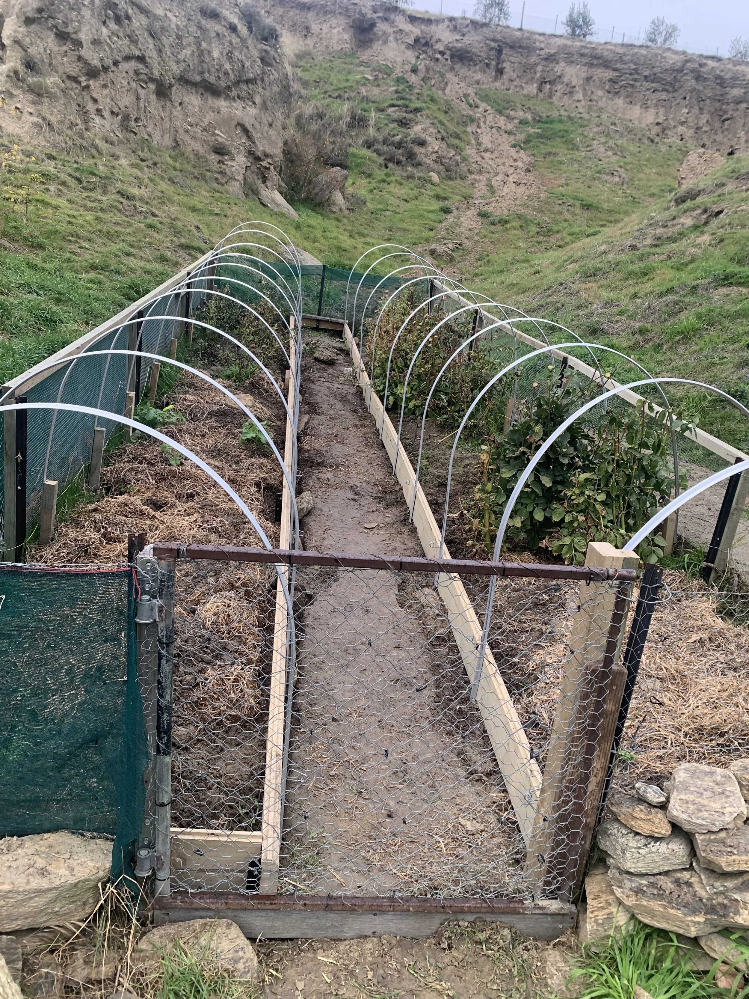 A small garden with a pathway surrounded by a wooden frame, enclosed by a low gate, situated on a hillside with a rocky, grassy terrain.