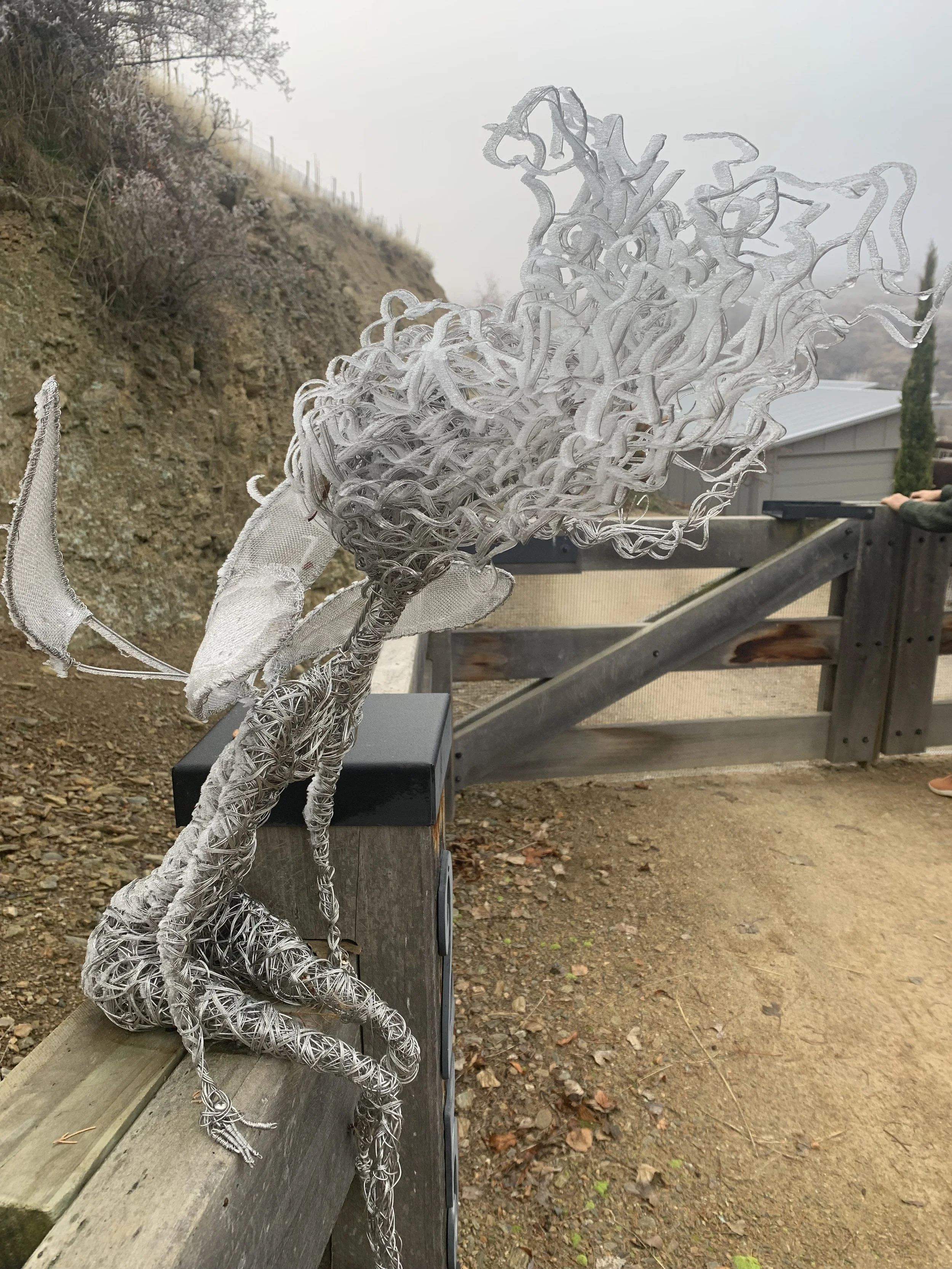 Wired metal sculpture of a dragon with wings, perched on a railing outdoors.