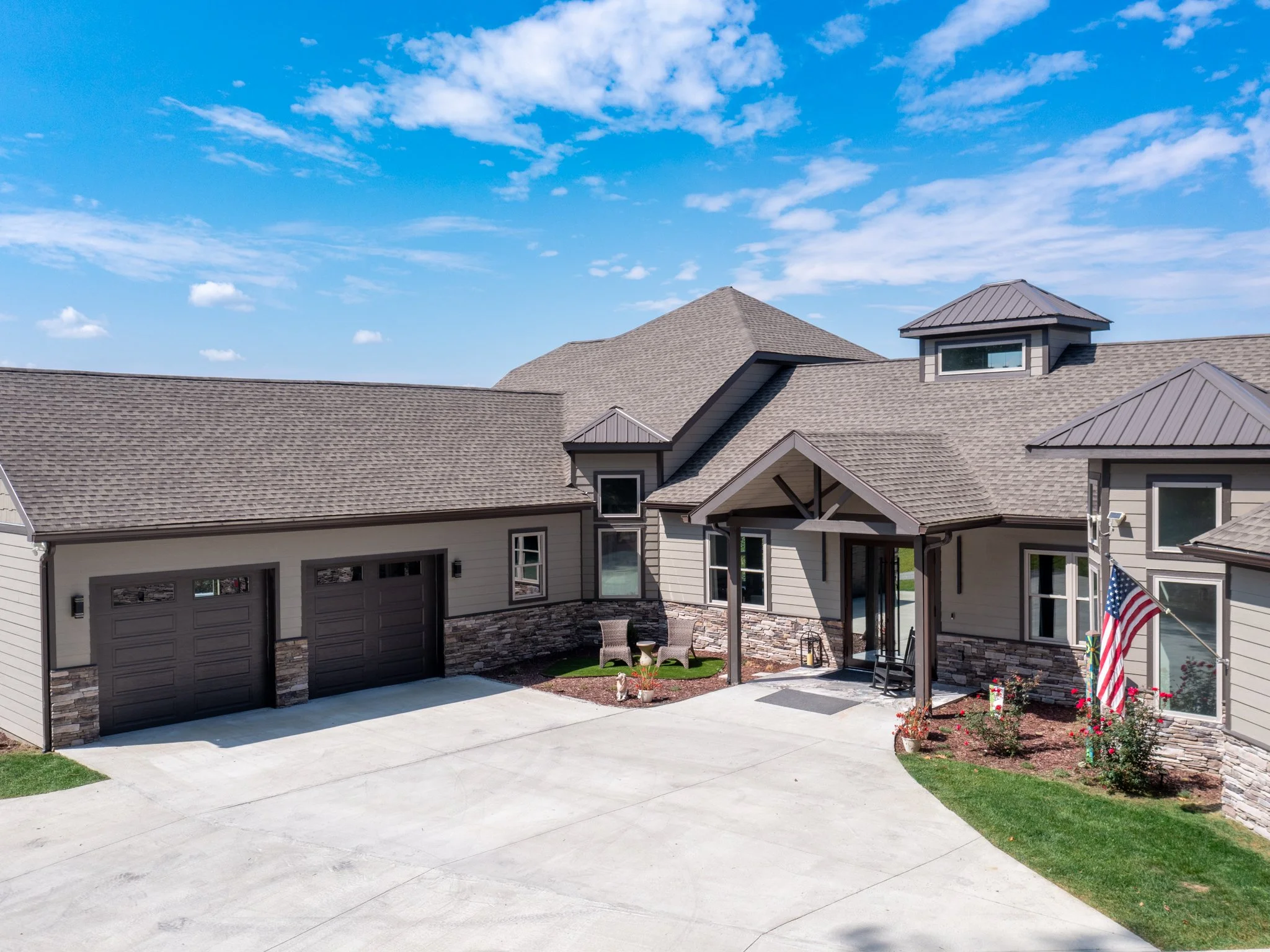 Front view of a modern house with a large driveway, three garage doors, a porch with a small seating area, and an American flag. The house has beige siding, stone accents, and multiple roof gables.