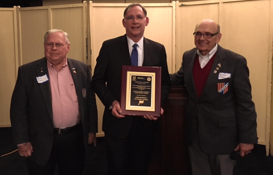 Three men in suits standing indoors, one holding a framed award or certificate. All are smiling, and the men on the left and right are wearing name tags and medals or ribbons.