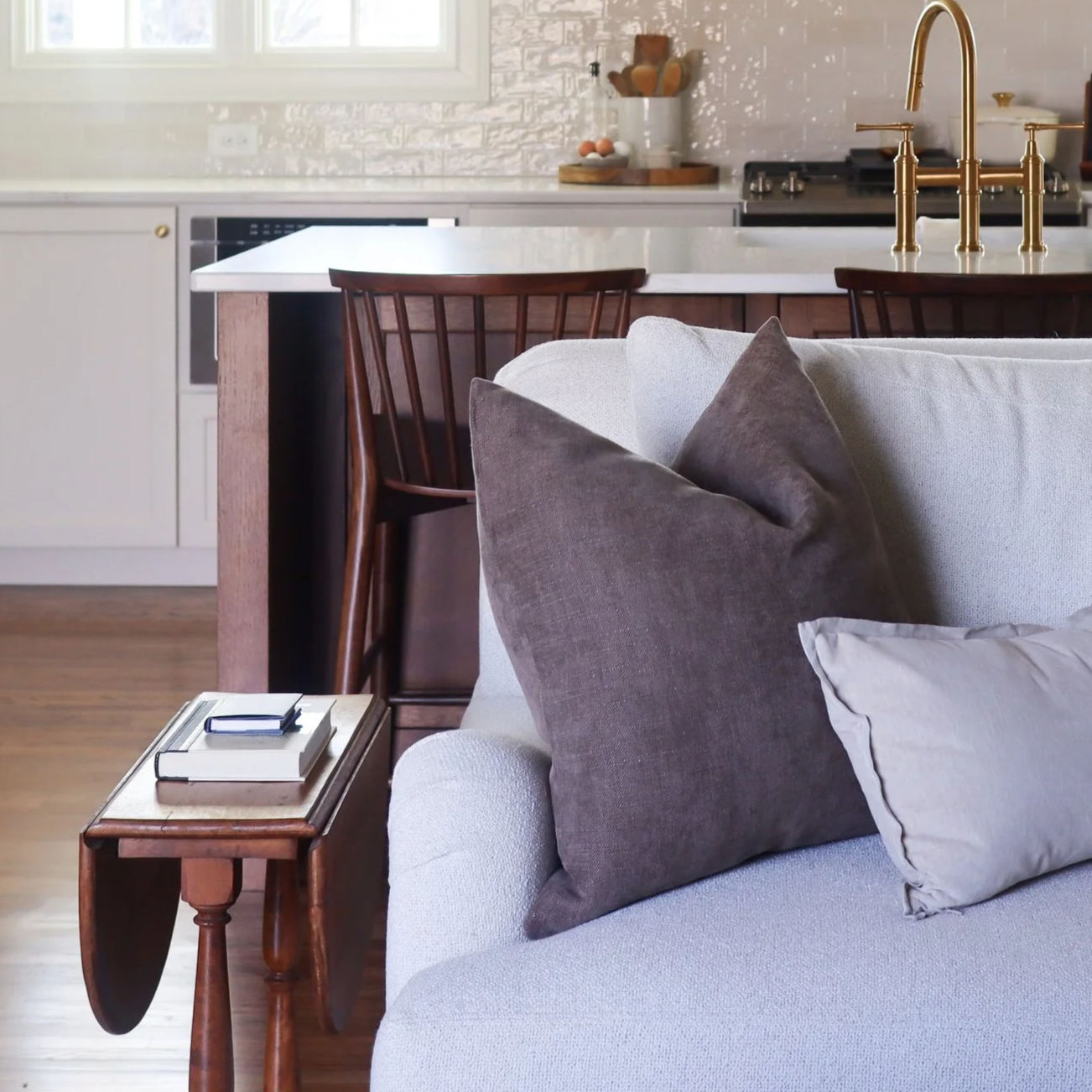 traditional neutral living room with cream couch, brown and tan pillows, wooden side table, kitchen with cream cabinets and wooden island in the background