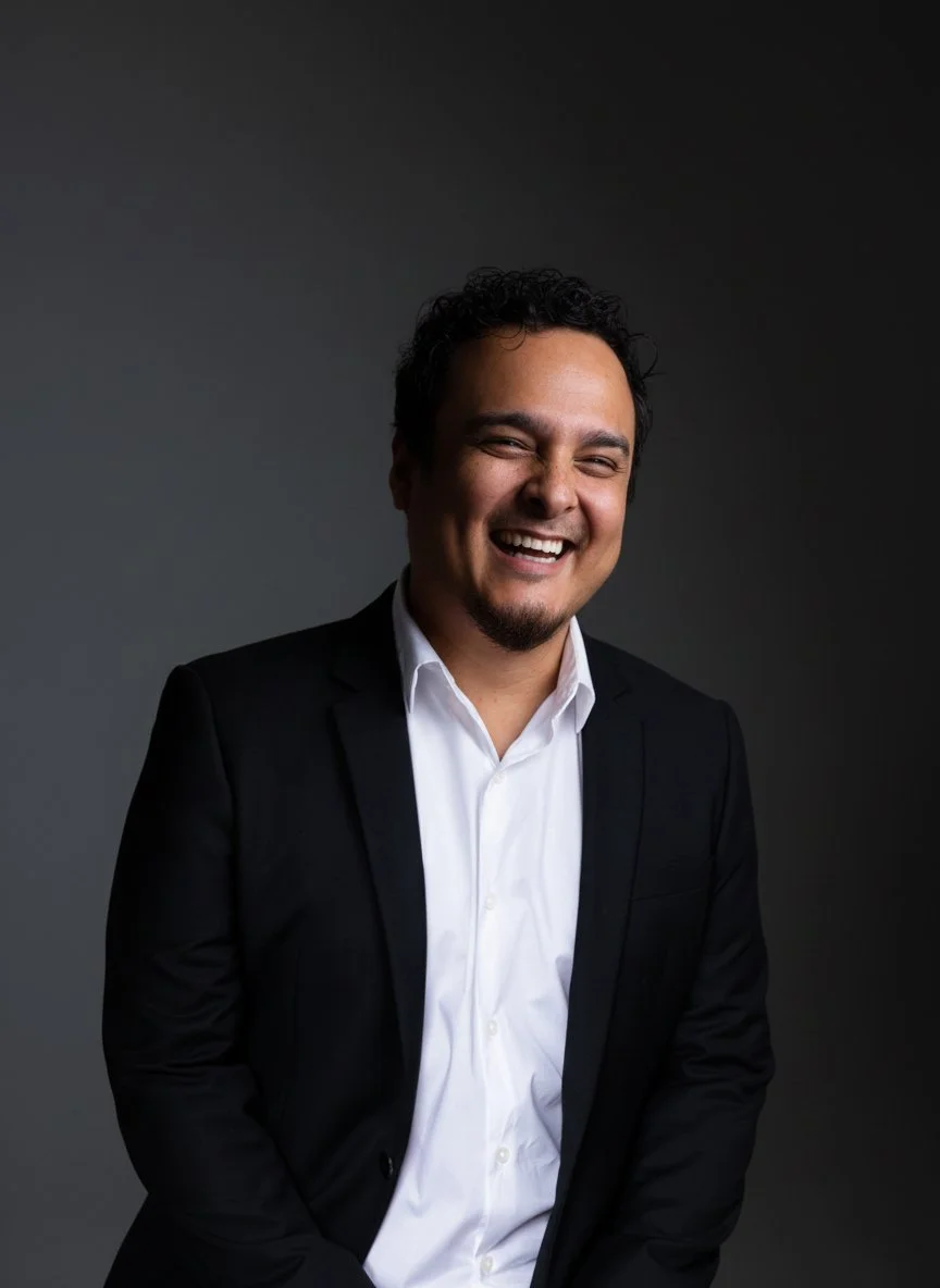 A man wearing a black suit and white shirt, smiling, against a plain white background.