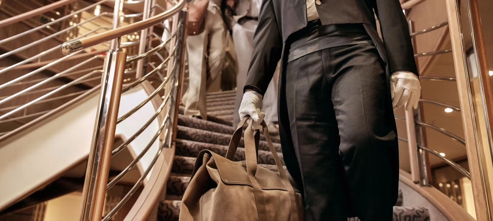 Person dressed in black and gray, wearing white gloves, carrying a beige duffel bag, ascending a staircase with a curved metal railing.