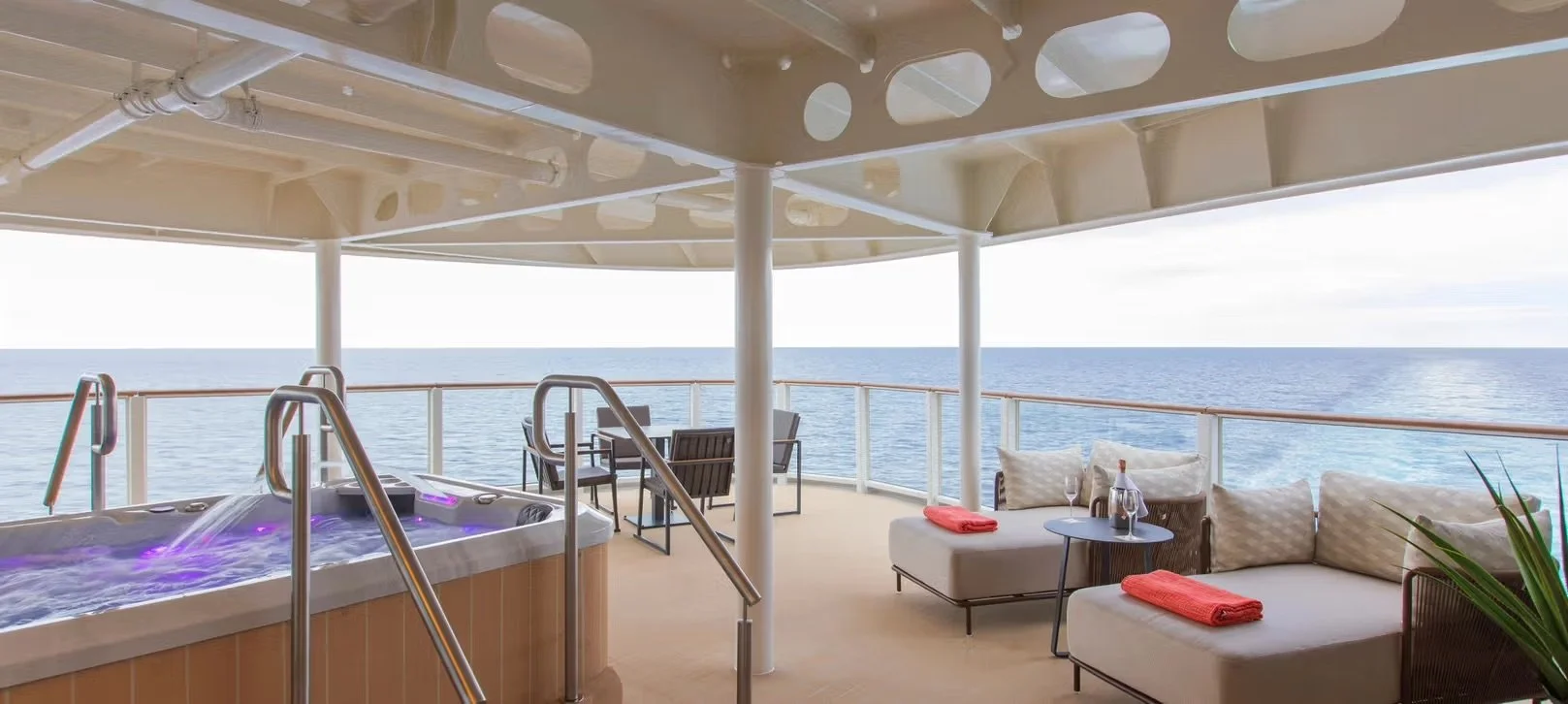 A luxurious outdoor deck on a cruise ship with a hot tub, lounge chairs, and seating area with pillows, overlooking the ocean.