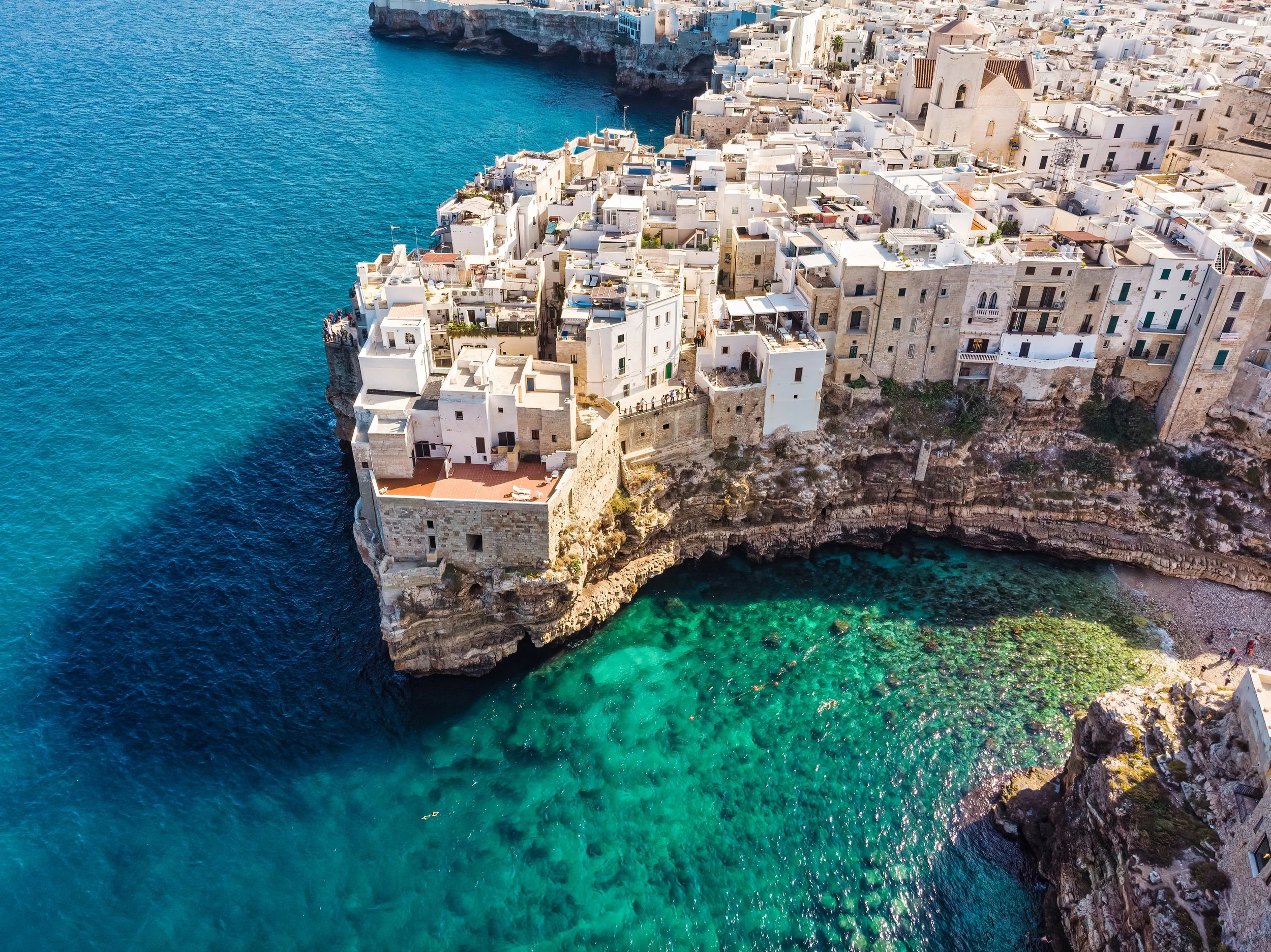 Aerial view of a coastal town with white buildings built on cliffs overlooking turquoise water, with some buildings on the edge of a rocky shoreline.